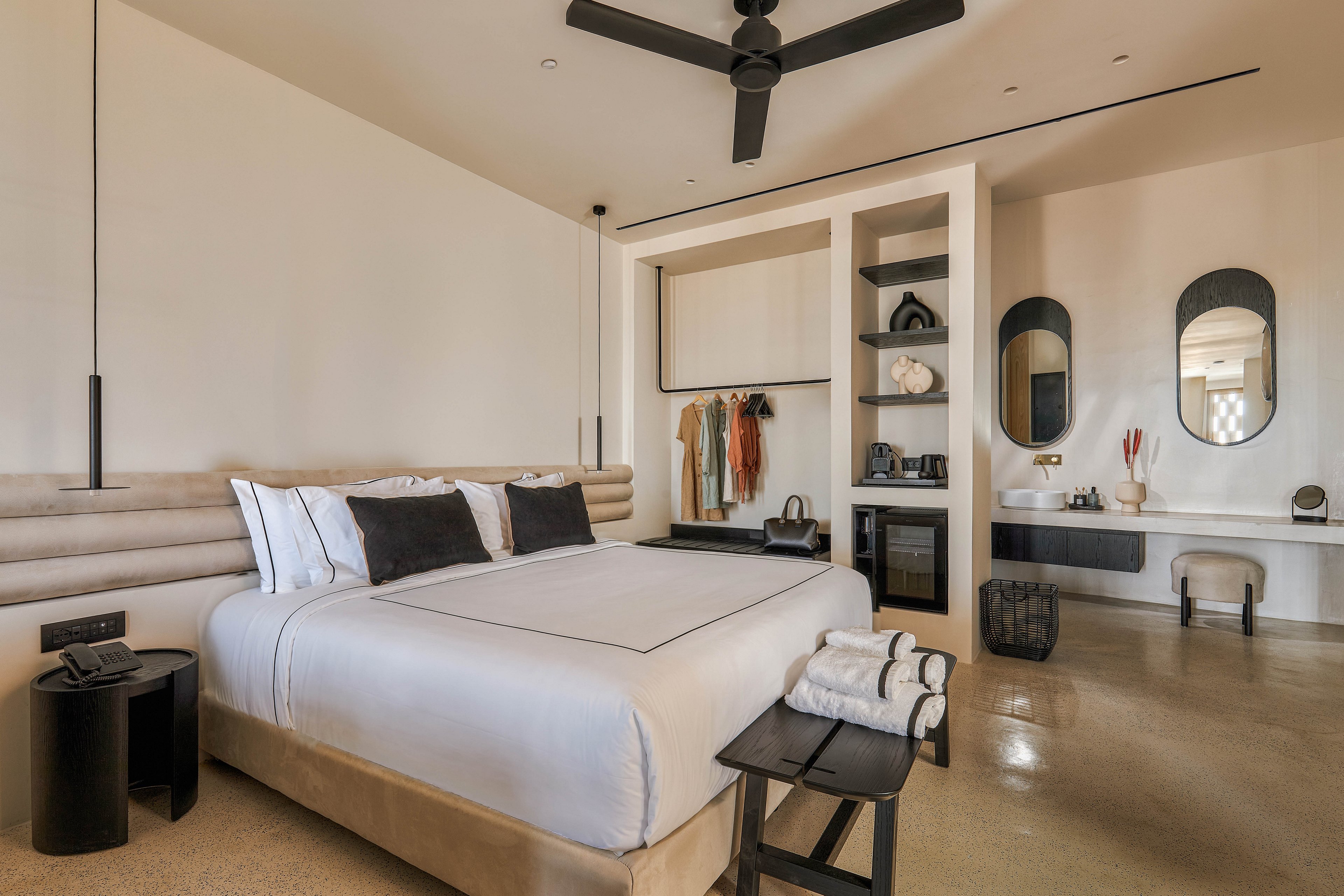 Contemporary hotel room featuring a large neatly made bed with white linens and black accent pillows. The space includes built-in open closets with hanging clothes and shelves holding decor items and a minibar. A sleek desk with twin oval mirrors and a stool sits along the wall, complemented by neutral tones and polished flooring. Black pendant lights, a ceiling fan, and simple accessories create a calm, minimalist atmosphere.