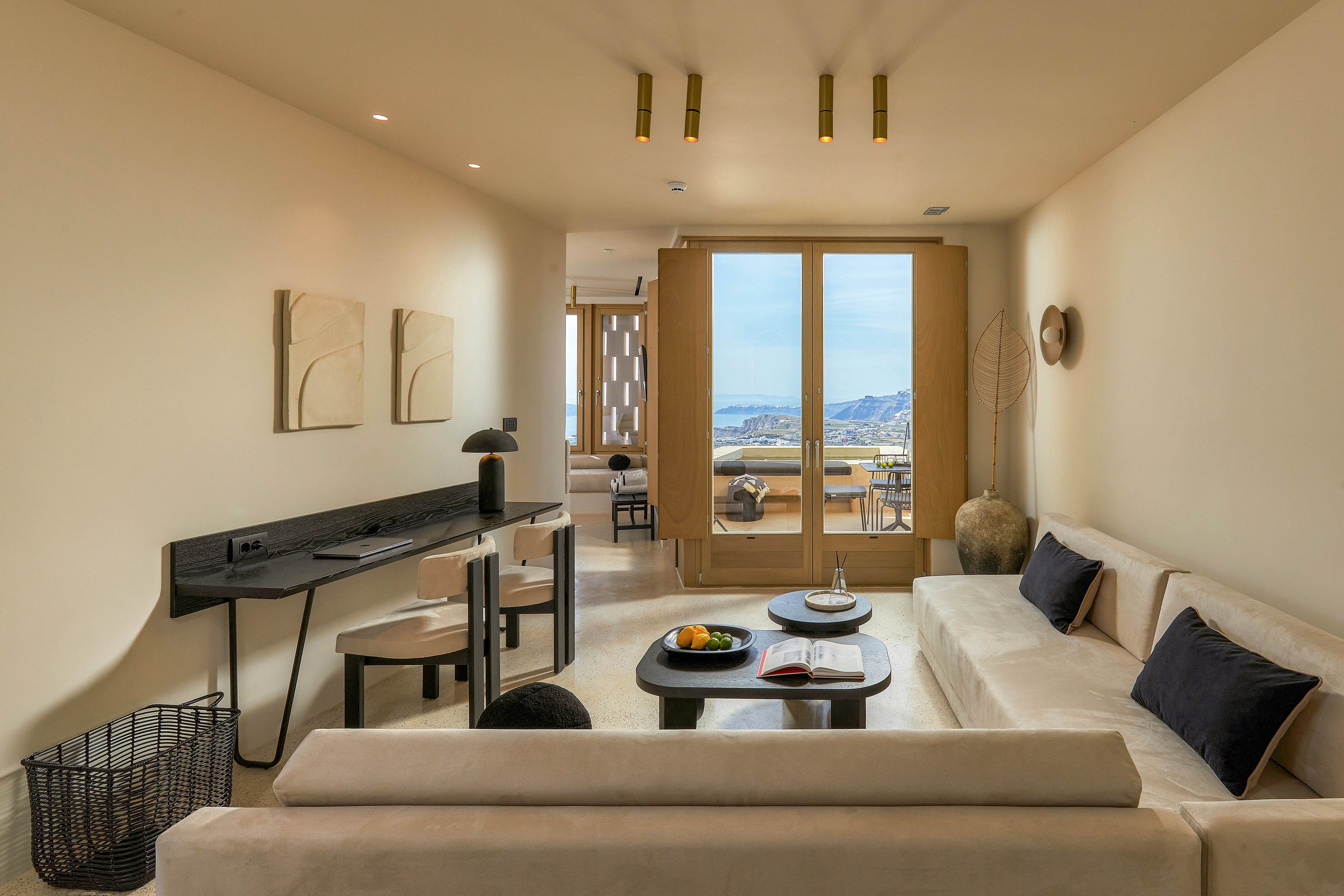 A contemporary hotel-style living room features a cream sofa, black accent pillows, and a sleek black desk along one wall. Minimalist artwork and recessed ceiling lights enhance the clean, modern design. Large glass doors open to a balcony with outdoor seating and a scenic view of mountains and water in the distance. A central coffee table with fruit and an open book adds a relaxed, inviting atmosphere.