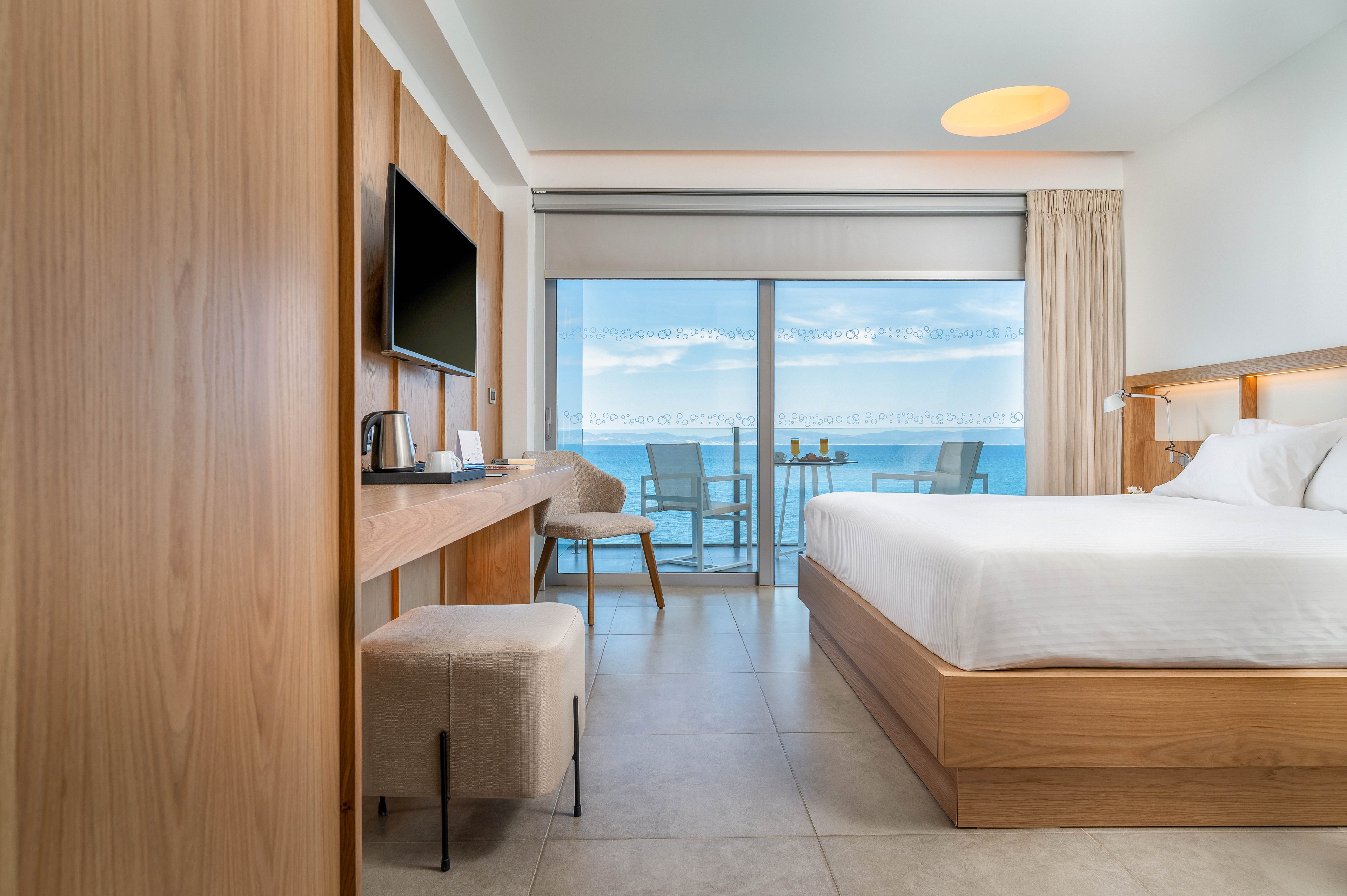 Bright contemporary hotel room features a large bed with white linens and light wood furniture. A wall-mounted television and long desk with a kettle and cups line one side of the room. Floor-to-ceiling glass doors open to a balcony with chairs and a table overlooking a calm ocean and blue sky. Neutral tones, clean lines, and soft lighting create a minimalist, relaxing atmosphere.