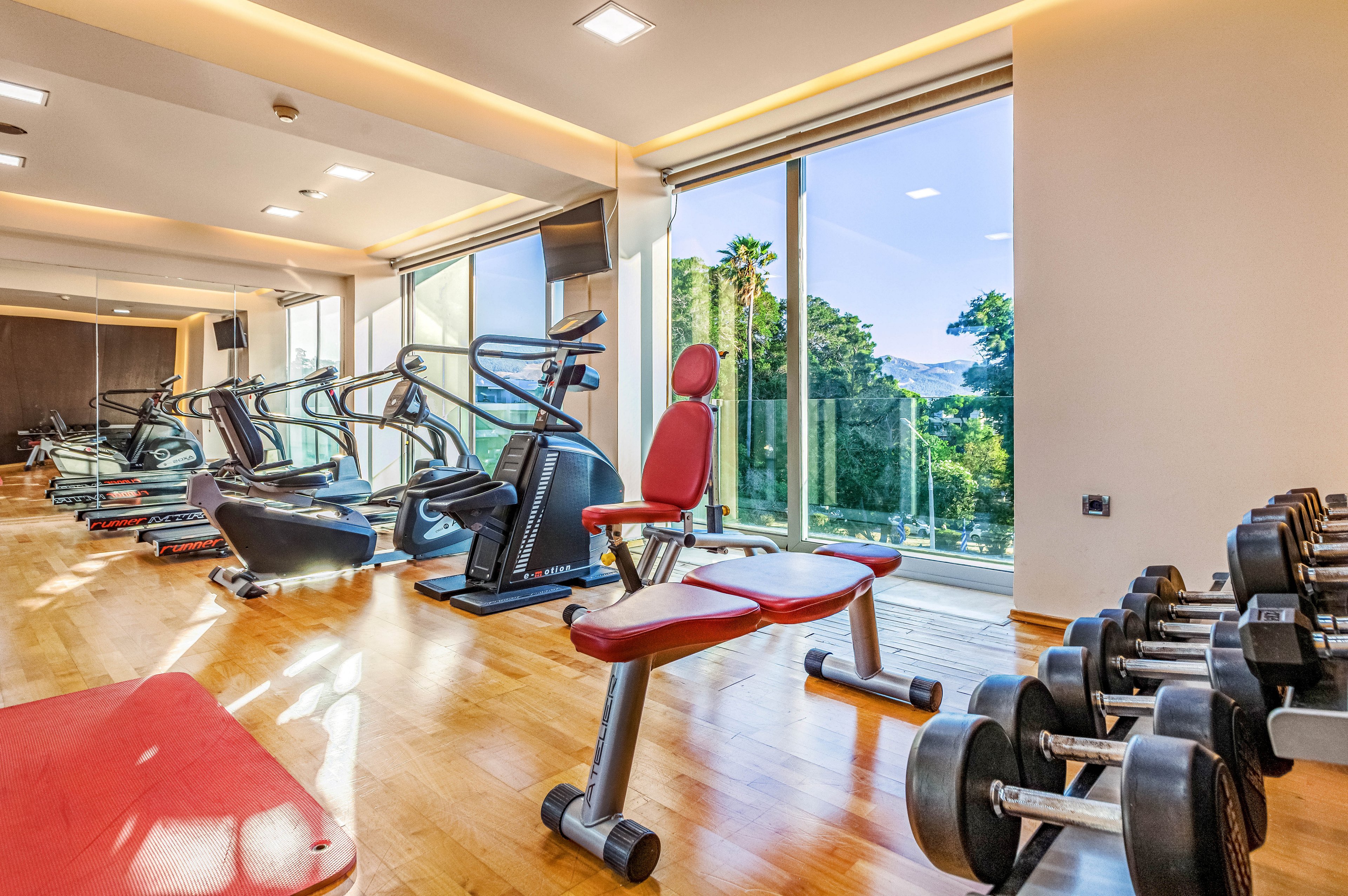 Bright, contemporary fitness room featuring treadmills, exercise bikes, and a red adjustable weight bench. Large floor-to-ceiling windows reveal a scenic outdoor view with trees and hills under a clear sky. Dumbbells are neatly arranged on a rack along the right side, and a wooden floor adds warmth to the clean, minimalist interior. The space appears empty, ready for individual workouts or personal training sessions.