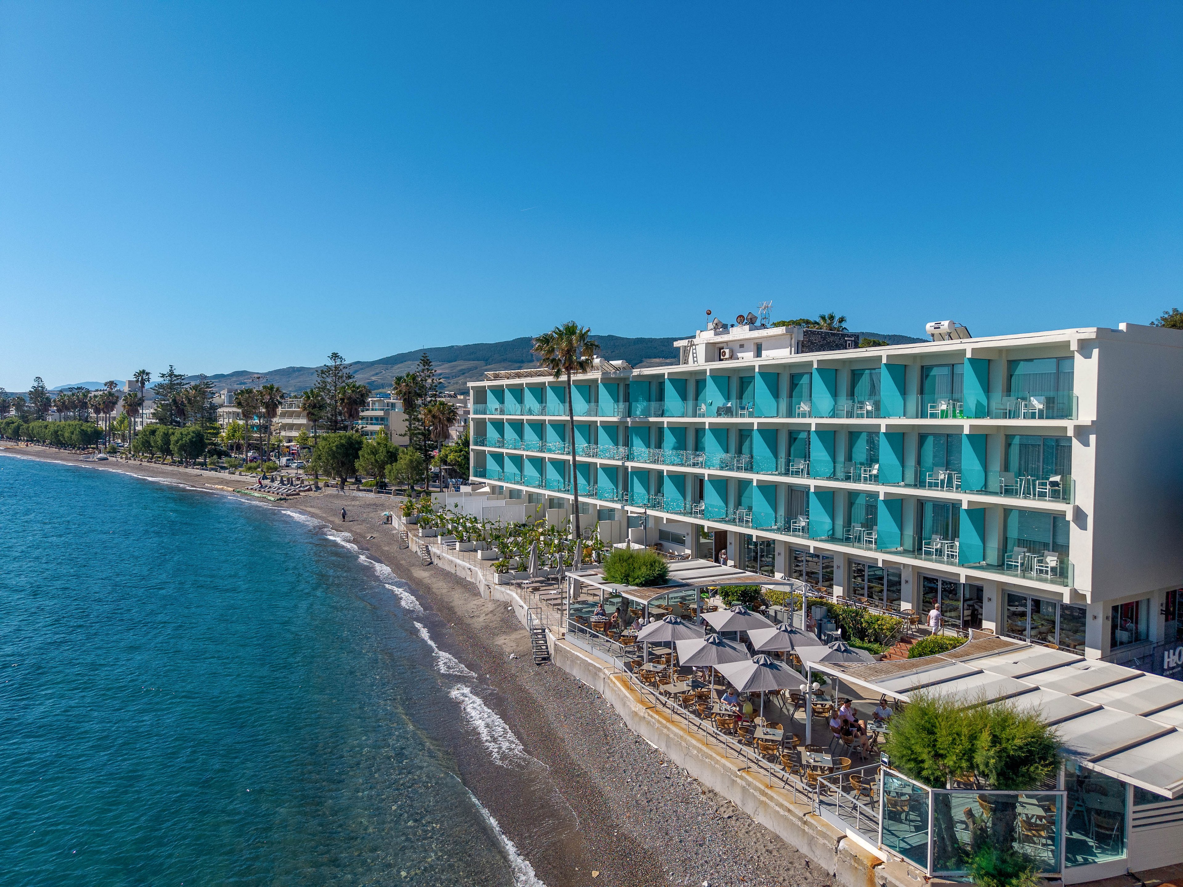 A contemporary multi-story hotel stands directly on a pebbled beachfront beside calm blue sea water. Turquoise balcony panels line the building, giving it a bright and modern appearance. Outdoor seating with umbrellas and greenery occupies the ground-level terrace overlooking the shore. In the background, coastal hills and palm trees complete the scenic seaside setting under a clear blue sky.