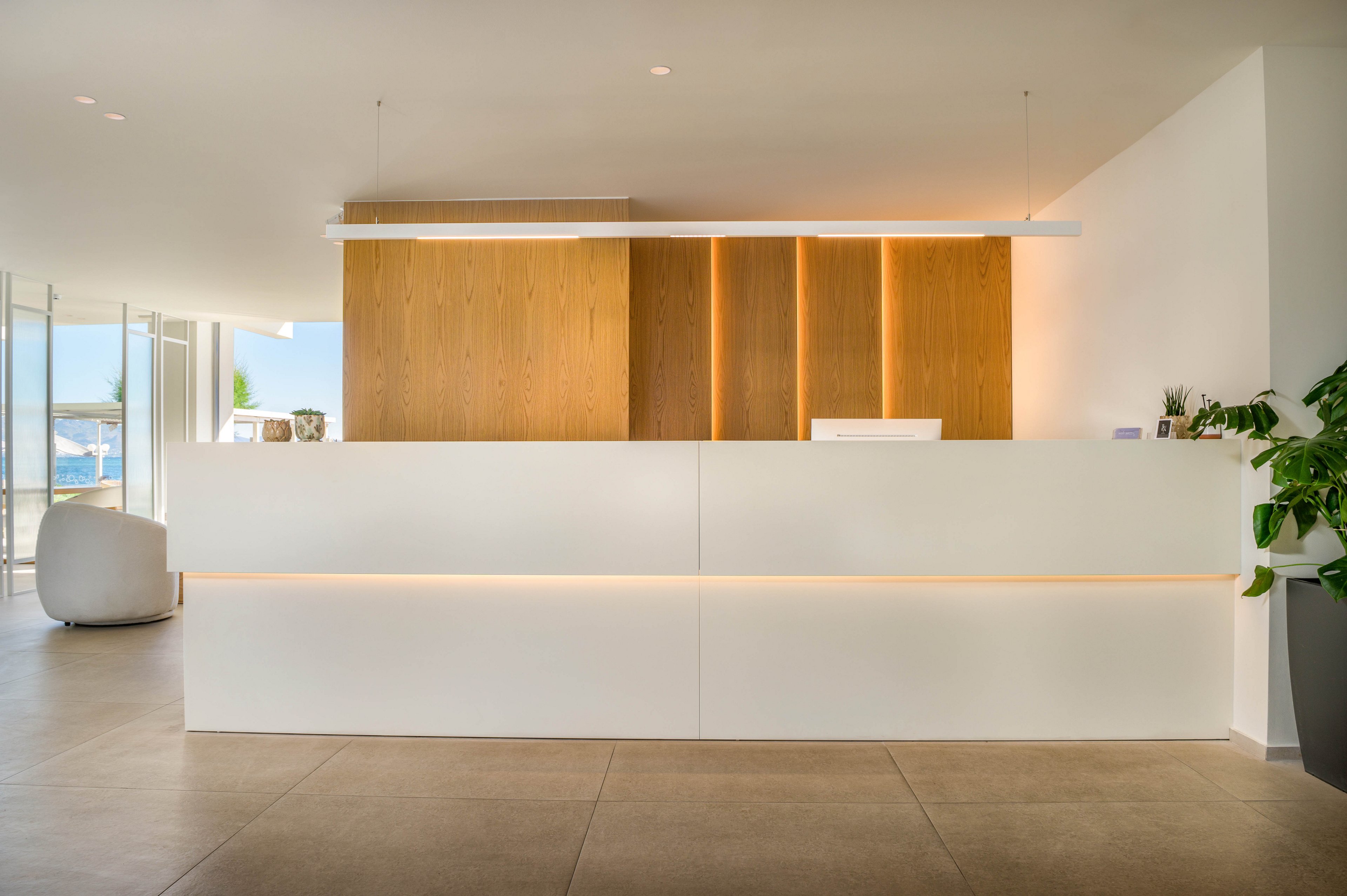Contemporary reception area featuring a large white front desk with integrated warm lighting. A wooden wall with vertical light strips forms a clean backdrop behind the counter. The space is bright and open, with neutral flooring and a white lounge chair visible to the side. Potted greenery and decorative objects add subtle accents to the minimalist design.