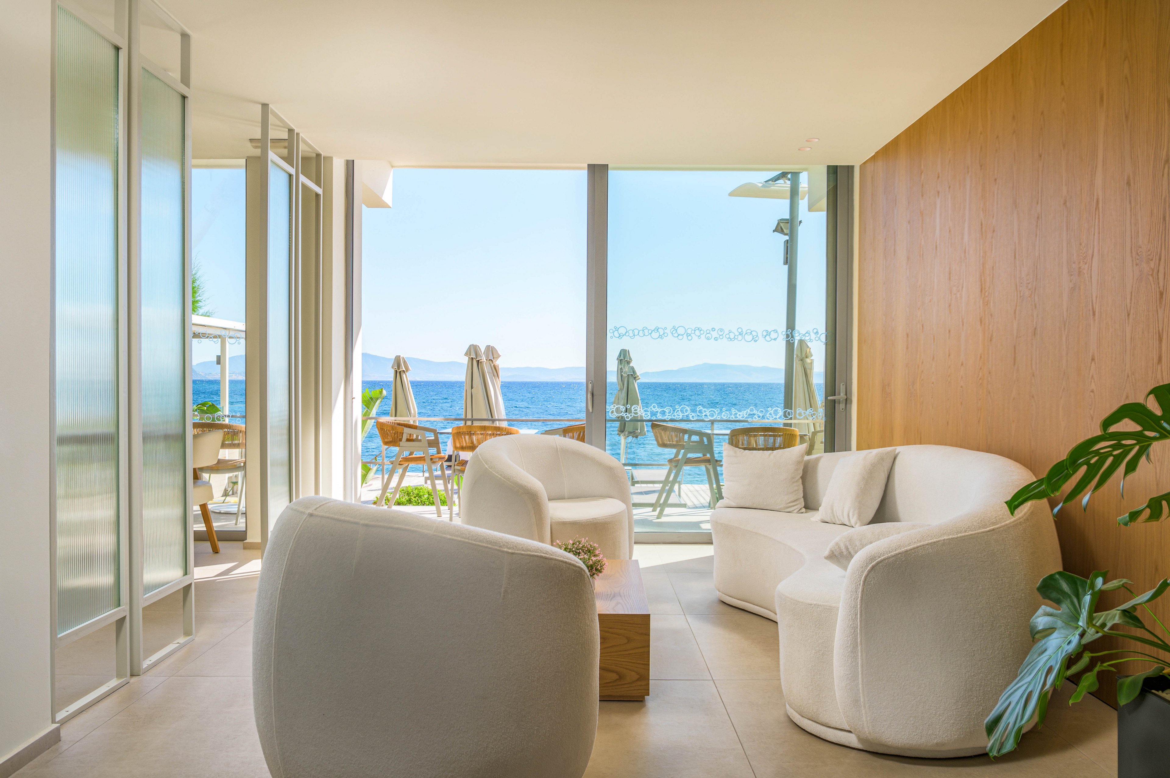 Bright contemporary lounge area features rounded white armchairs arranged around a small wooden table. Floor-to-ceiling glass doors open to a terrace with sun loungers and umbrellas overlooking the sea. Natural light floods the minimalist interior, highlighting clean lines and neutral tones. A green potted plant adds a touch of nature to the calm, coastal setting.