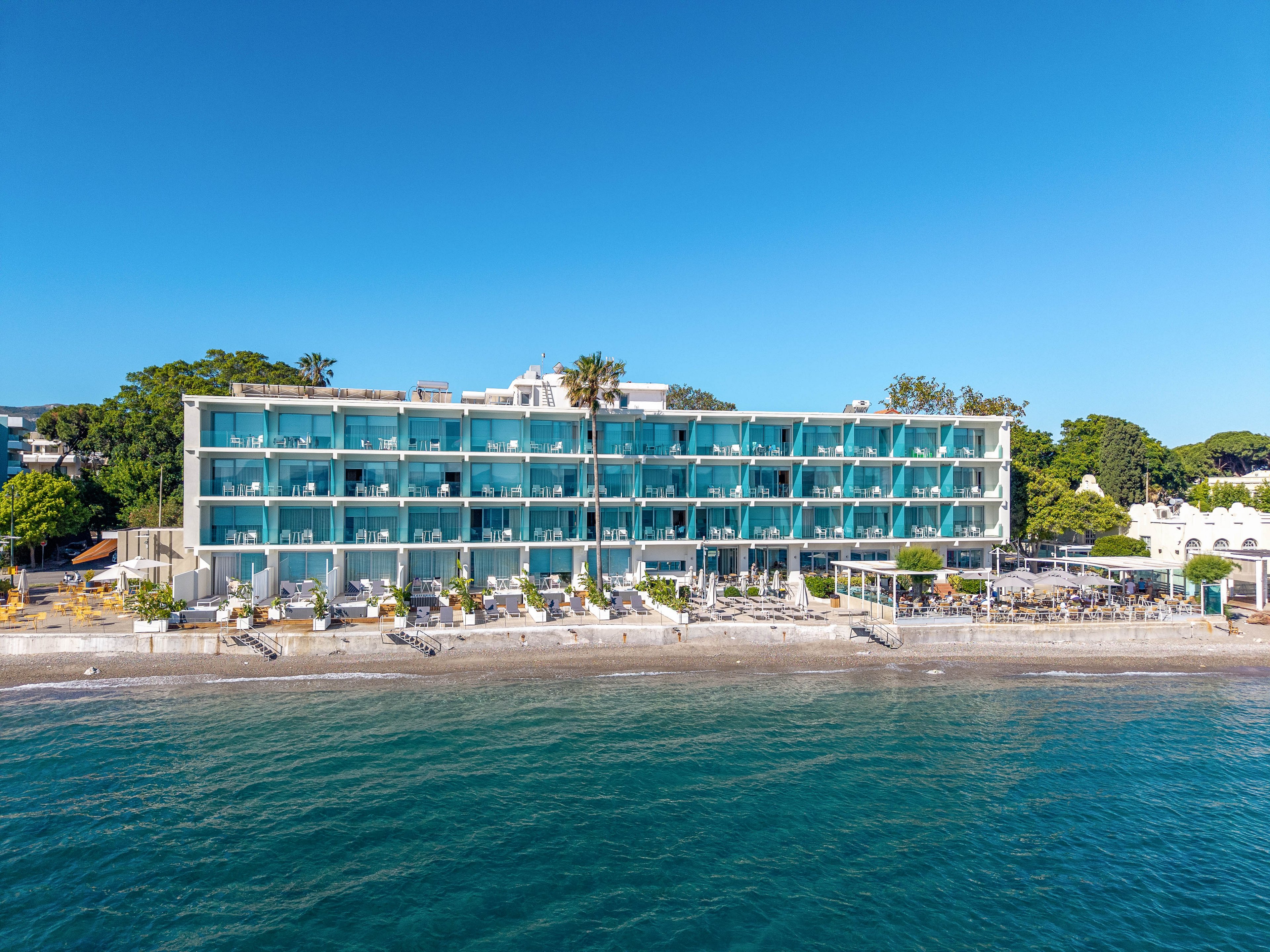A contemporary multi-story hotel stands directly on a sandy beachfront, featuring rows of glass-fronted balconies facing the sea. Sun loungers and umbrellas are arranged along the shore, emphasizing a relaxing vacation setting. Calm blue water fills the foreground, while clear skies and surrounding greenery complete the coastal scene. The overall atmosphere is bright, inviting, and ideal for seaside holidays.