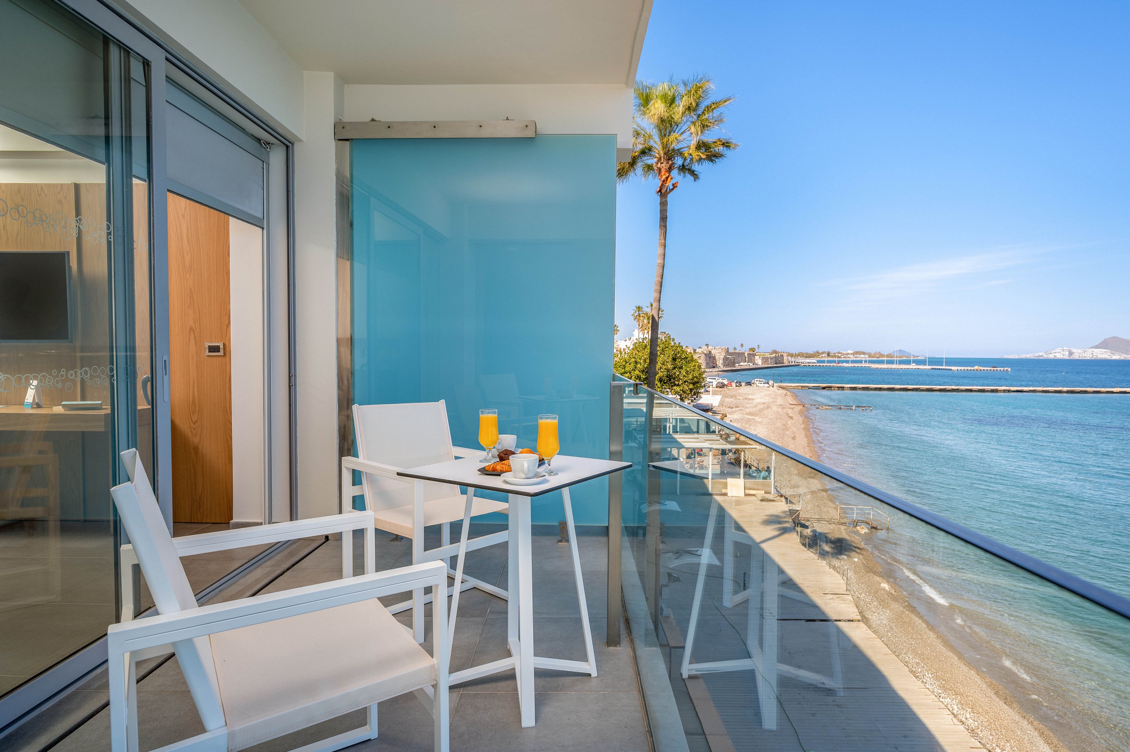 A modern hotel balcony features white chairs and a small table set with orange juice and a light snack. Glass railings frame a clear view of the sandy beach and calm blue sea below. A tall palm tree and distant buildings line the coastline under a bright, cloudless sky. The scene evokes a relaxed vacation atmosphere with clean, contemporary design and vibrant coastal colors.