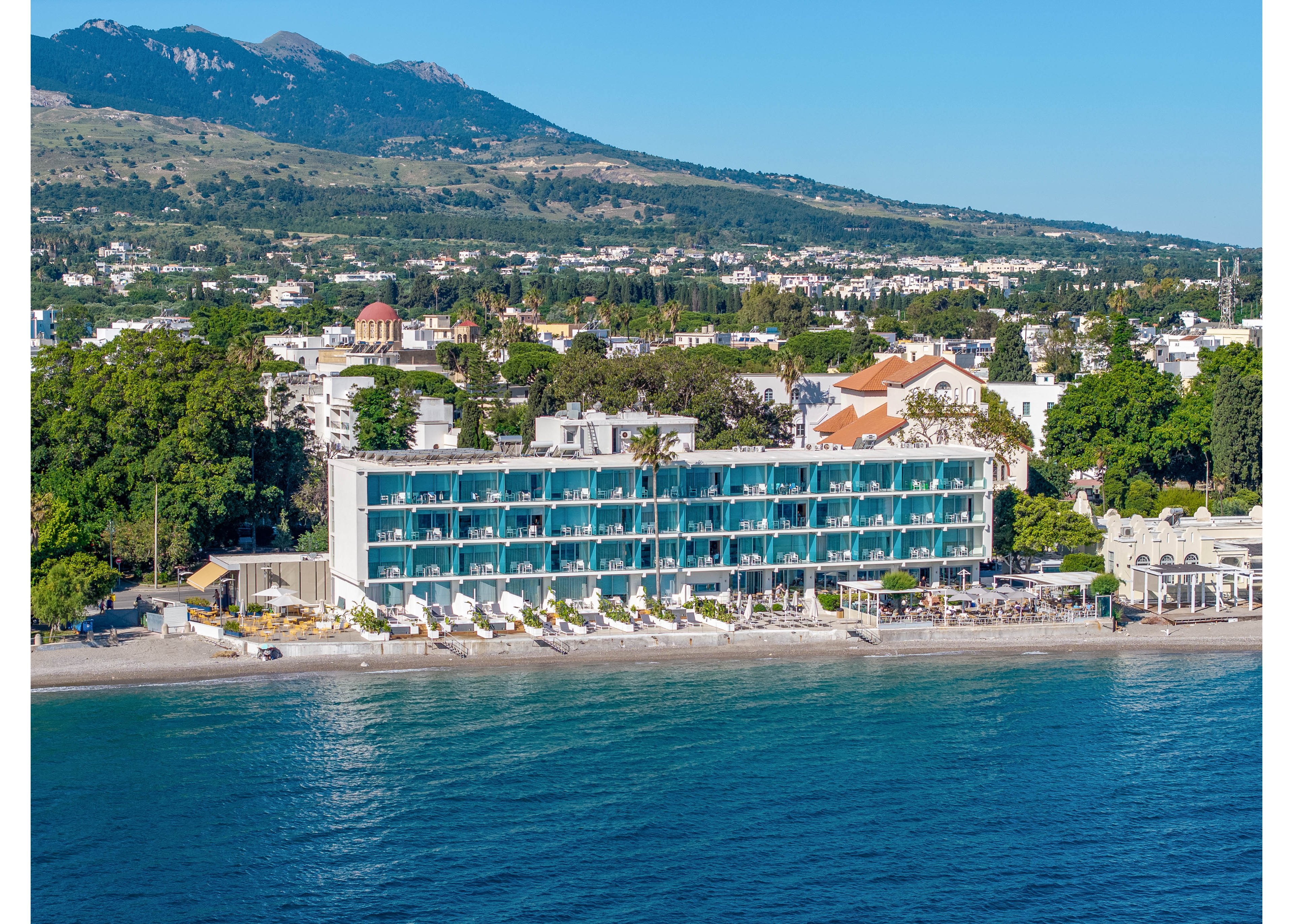 A contemporary multi-story hotel stands directly on a calm beachfront, with balconies facing the sea. Sun loungers and umbrellas line the shore, emphasizing a relaxing vacation setting. Behind the hotel, lush greenery and scattered buildings extend toward a mountainous landscape under a clear blue sky. The overall scene conveys a peaceful coastal resort atmosphere with scenic natural surroundings.