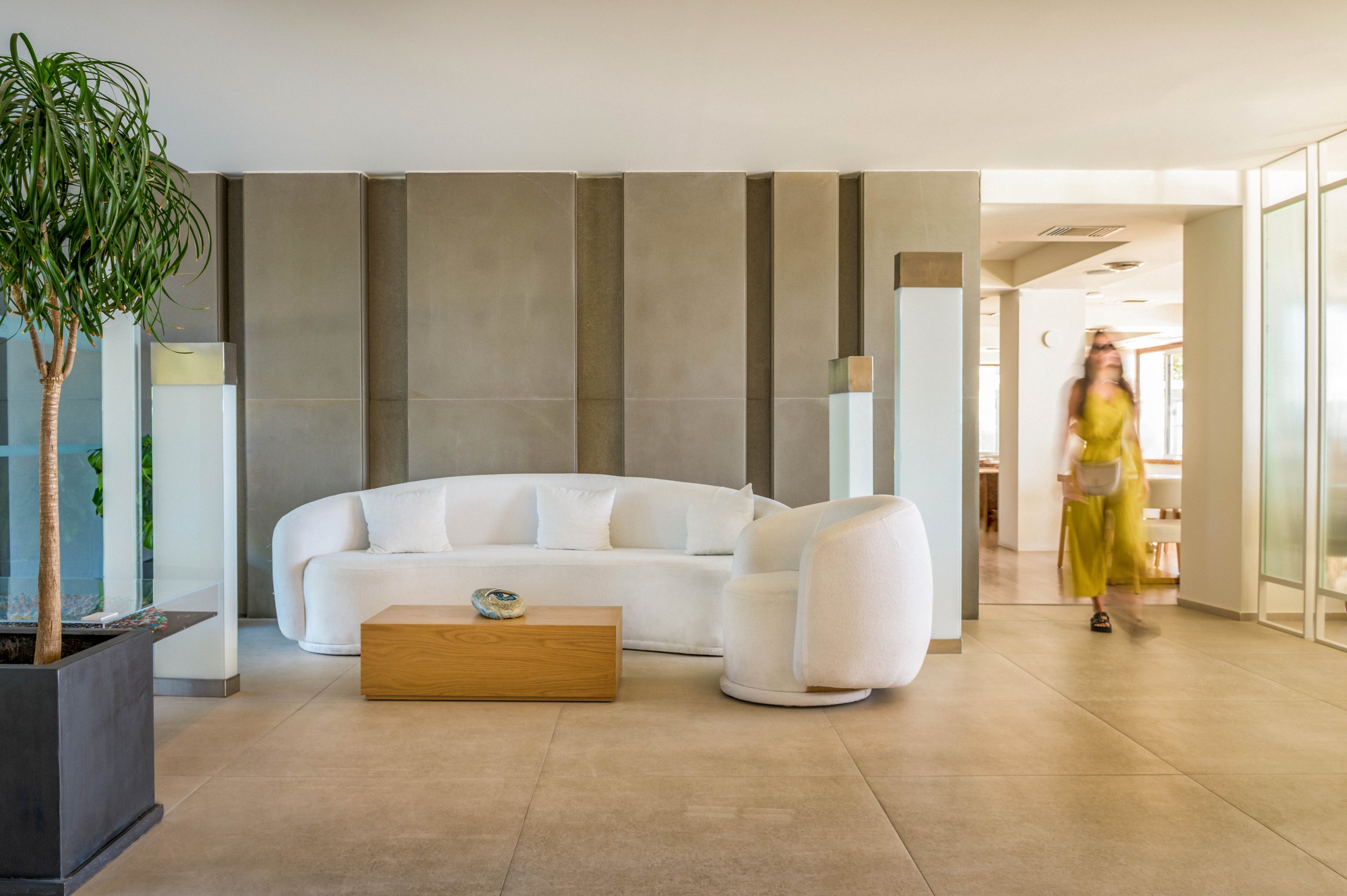 A contemporary lobby features a curved white sofa and matching armchair against a neutral wall. A low wooden coffee table sits in front of the seating area, holding a decorative object. Large floor tiles and glass partitions create a clean, open atmosphere. A potted plant and a blurred woman walking in a yellow dress add a touch of color and movement to the minimalist interior.