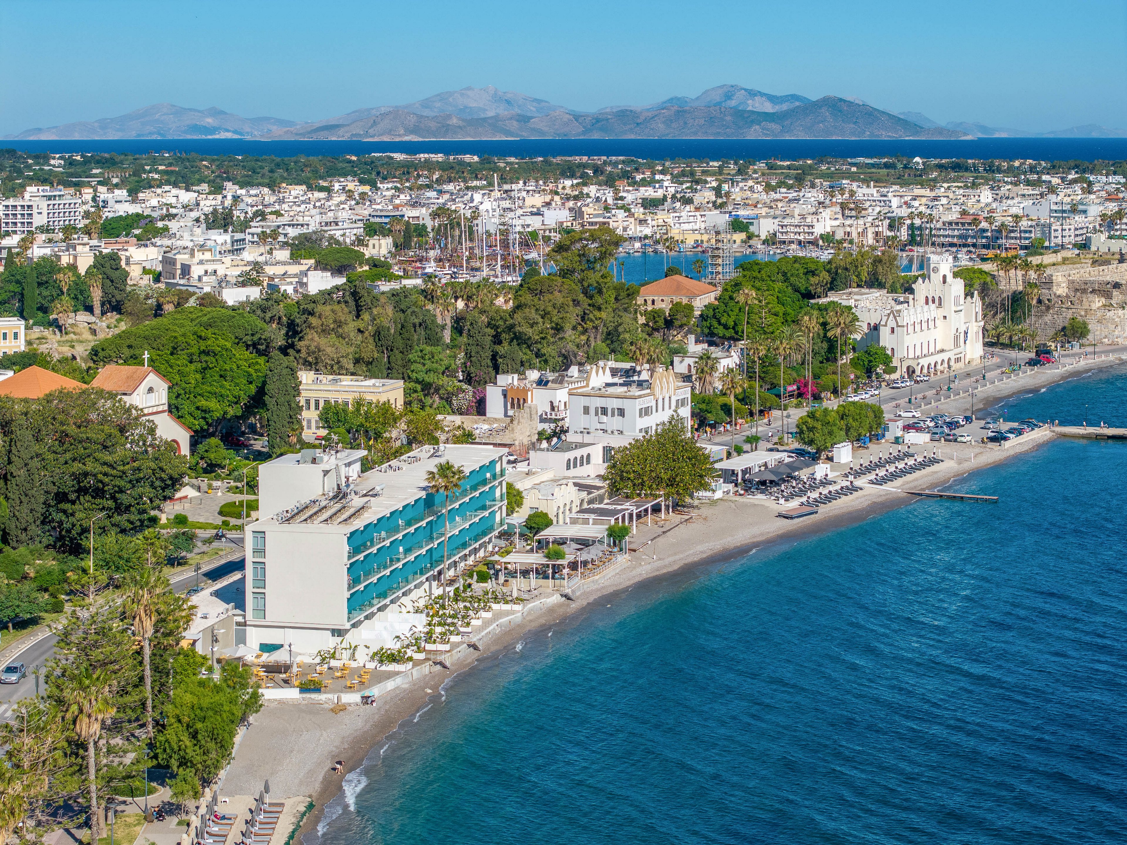 Aerial view of a coastal city shows a long beachfront promenade lined with modern and historic buildings beside clear blue sea water. Sun loungers and umbrellas are arranged along the narrow sandy beach, while a curving road follows the shoreline with parked cars and light traffic. In the distance, a marina with numerous masts and a dense urban area stretches toward hazy mountains under a bright blue sky. The scene suggests a Mediterranean-style resort destination with mixed architecture and vibrant waterfront activity.