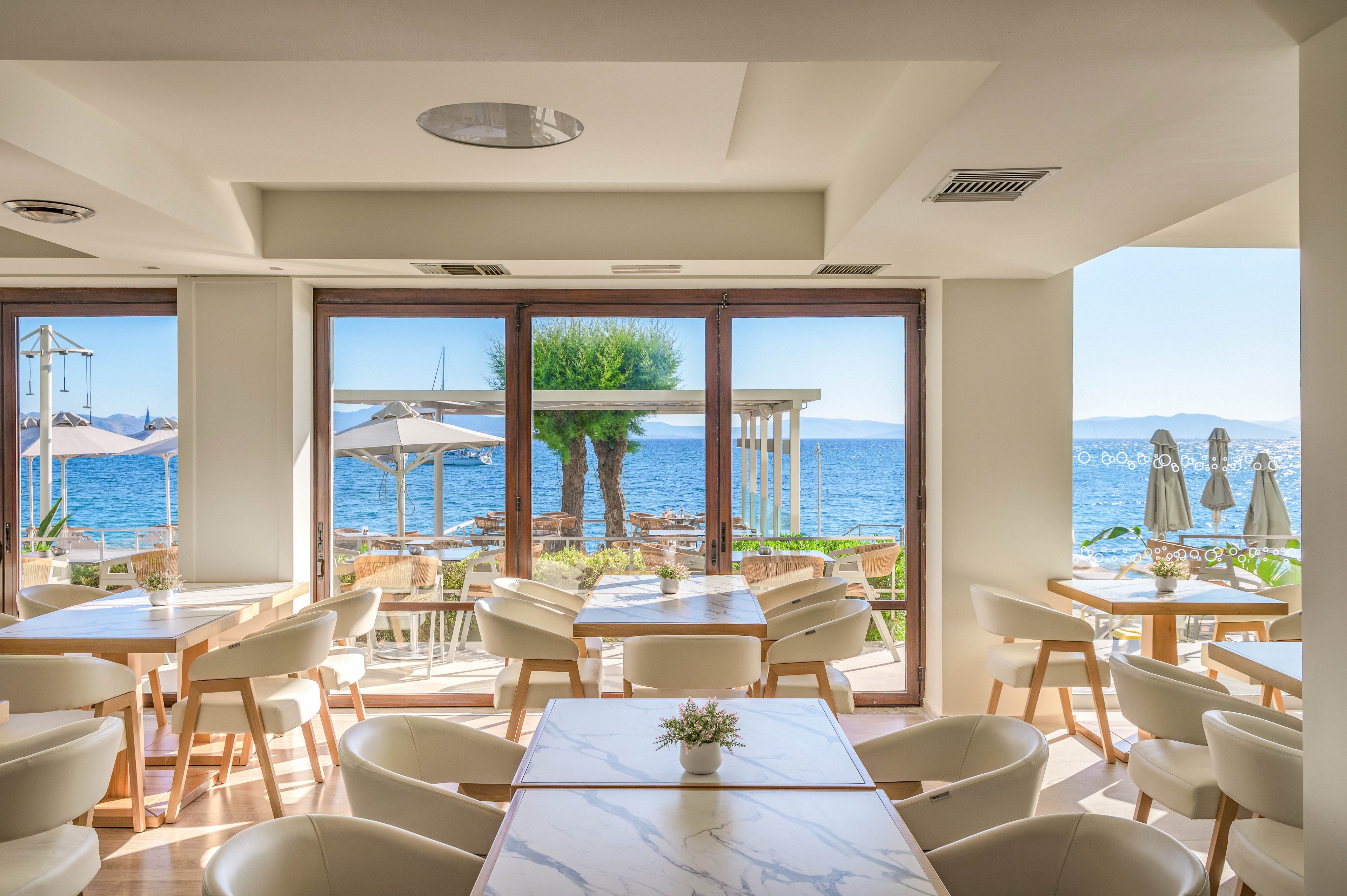 A modern, sunlit restaurant interior opens onto a terrace overlooking a calm blue sea. Light wooden tables and white upholstered chairs create a clean, contemporary dining space. Large windows frame outdoor seating with umbrellas, greenery, and distant boats on the water. The overall atmosphere is relaxed, airy, and inviting, ideal for coastal dining.