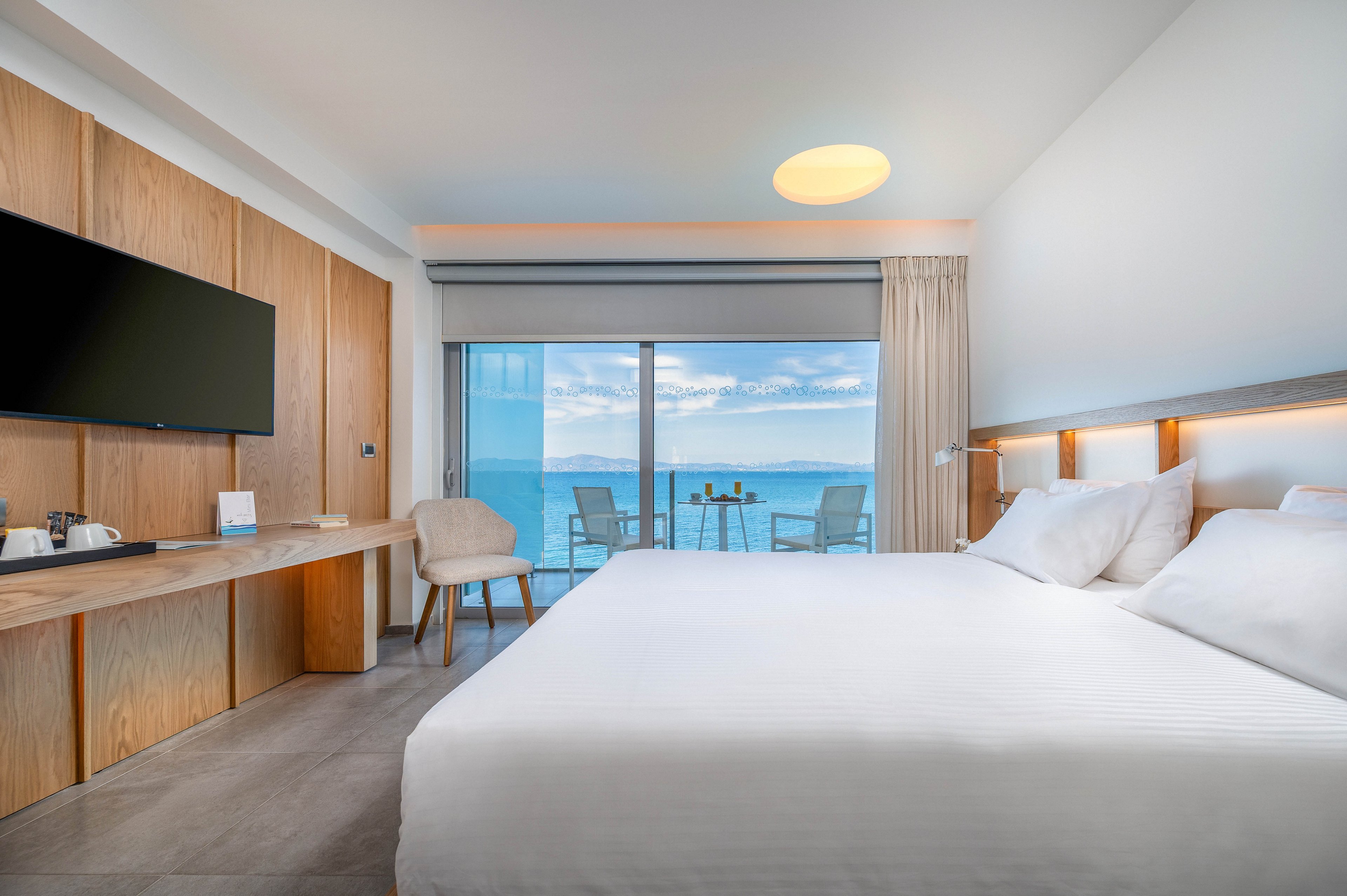Bright contemporary hotel room featuring a large neatly made bed and minimalist wooden furnishings. A flat-screen TV, desk area, and chair line one wall, with a small coffee and tea station visible. Floor-to-ceiling glass doors open onto a balcony with chairs and a table overlooking a calm blue ocean and sky. Soft neutral tones and clean lines create a relaxing, modern vacation atmosphere.