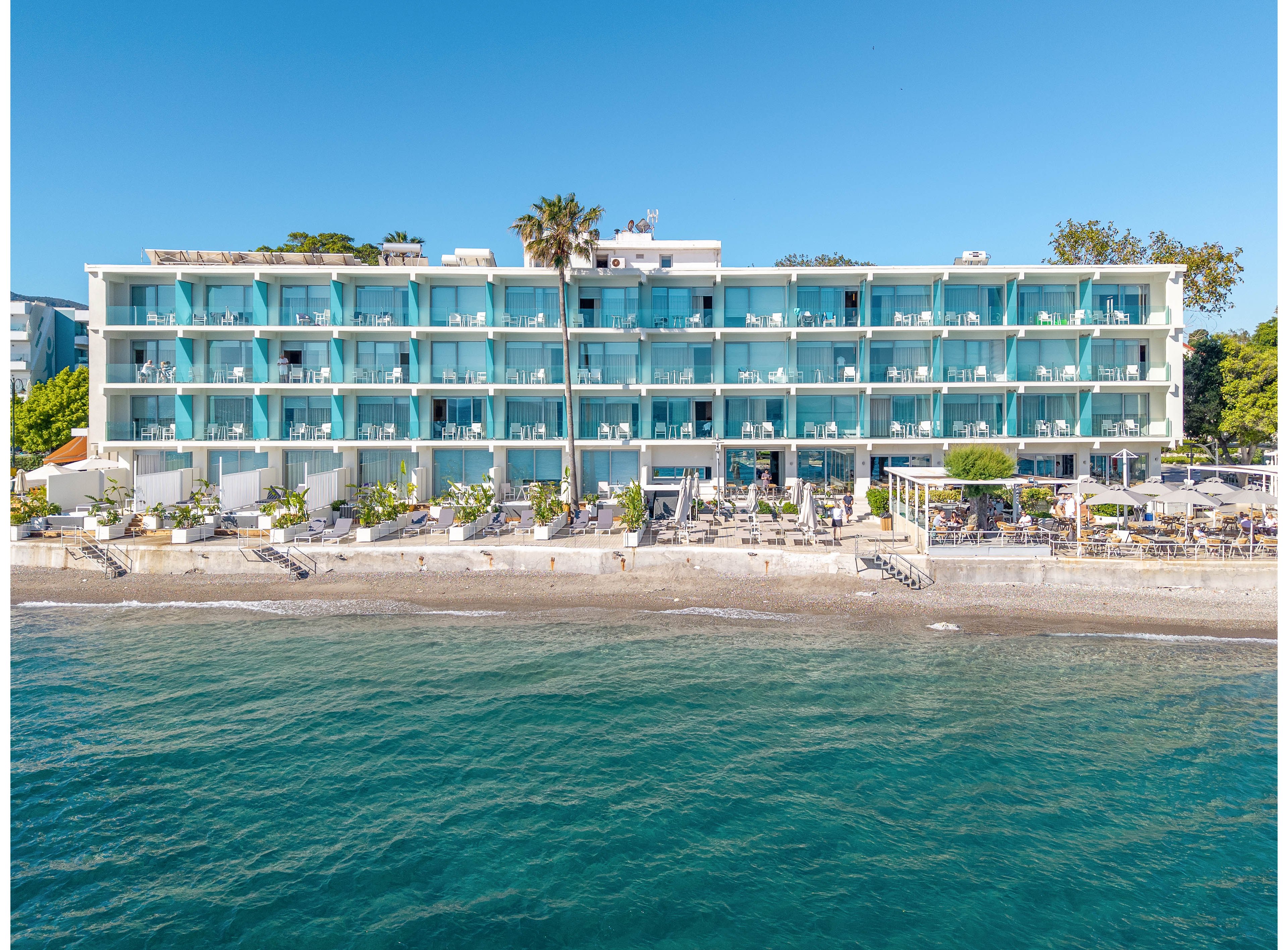 A contemporary multi-story hotel stands directly on a sandy beachfront, featuring rows of turquoise glass balconies facing the sea. Sun loungers and umbrellas line the shore, creating a relaxed holiday atmosphere by the water. The calm blue ocean occupies the foreground, while clear skies and surrounding greenery frame the building. The scene suggests a sunny coastal resort ideal for seaside vacations and leisure.