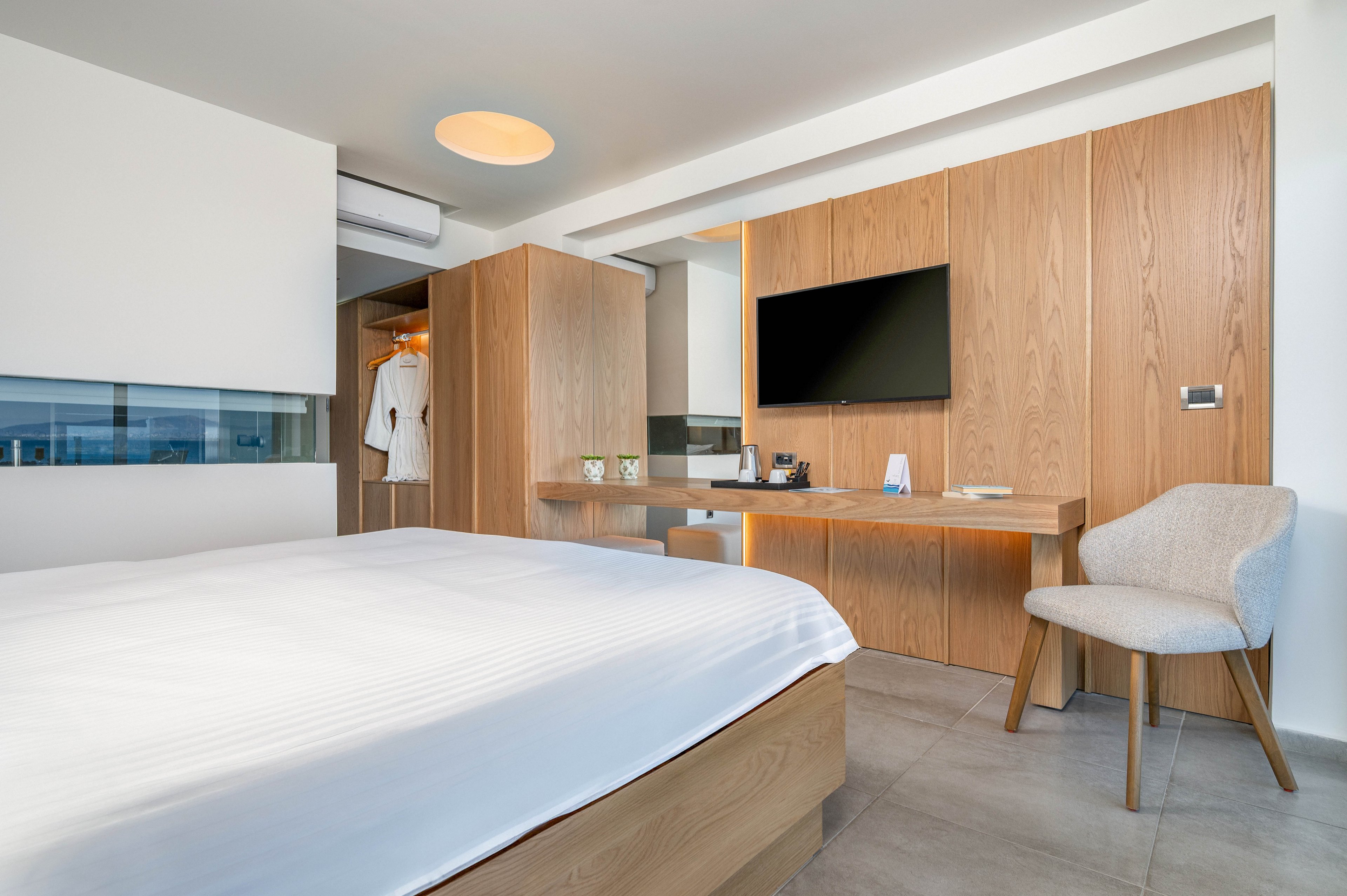 Contemporary hotel room featuring a large neatly made bed with white linens in the foreground. A wall-mounted flat-screen TV, wooden wardrobe, and minimalist desk create a clean, functional workspace. An upholstered chair and open closet with hanging clothes add comfort and practicality. Neutral tones, natural wood, and soft lighting give the space a calm, modern atmosphere.