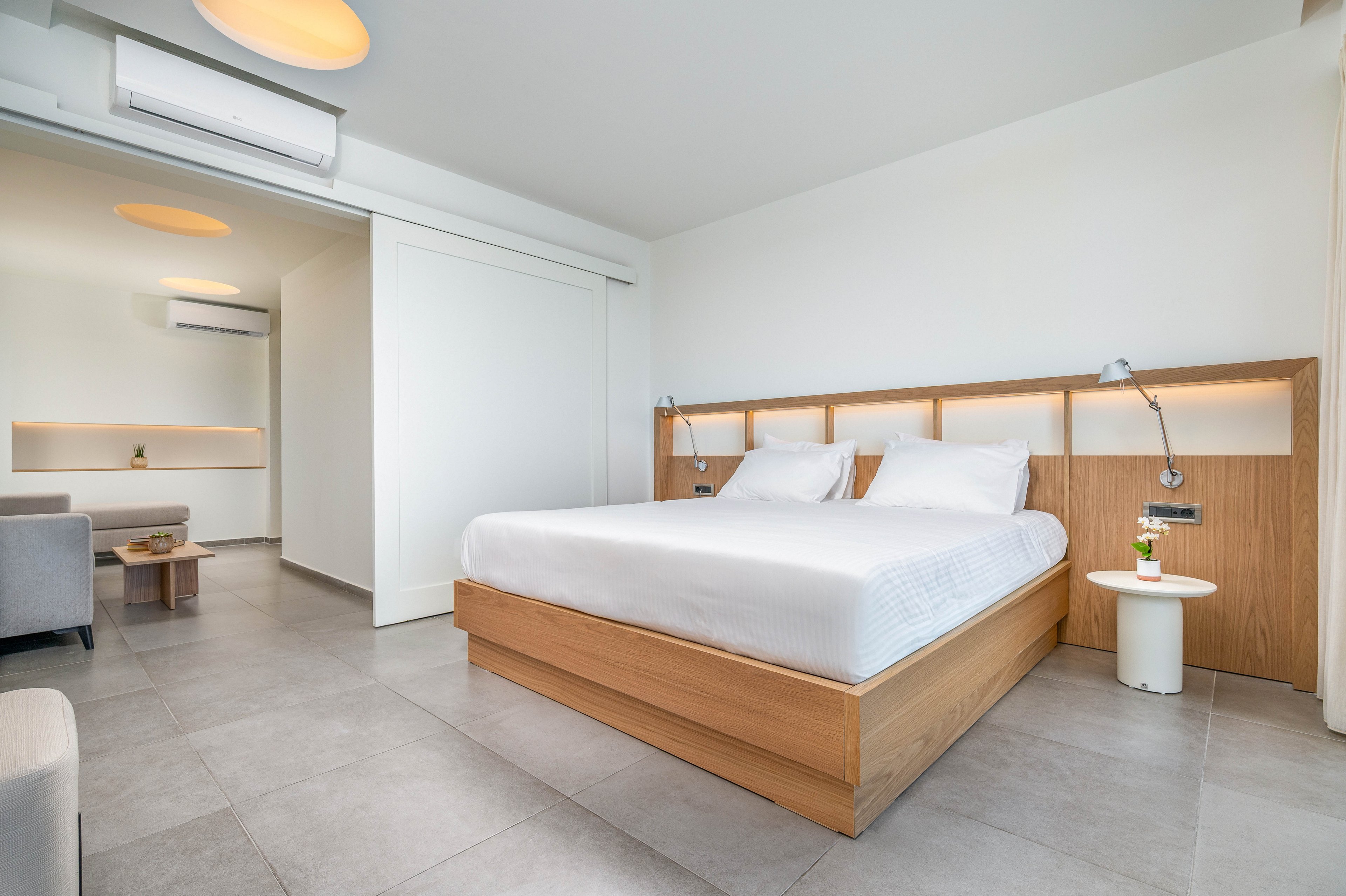 A spacious modern bedroom features a large double bed with a wooden frame and crisp white linens. The room has light gray tiled flooring, white walls, and built-in wooden shelving with integrated lighting. A small round side table with a drink adds a touch of color near the bed, while a sitting area is visible in the background. Overall, the space feels clean, bright, and minimalist, emphasizing comfort and contemporary design.