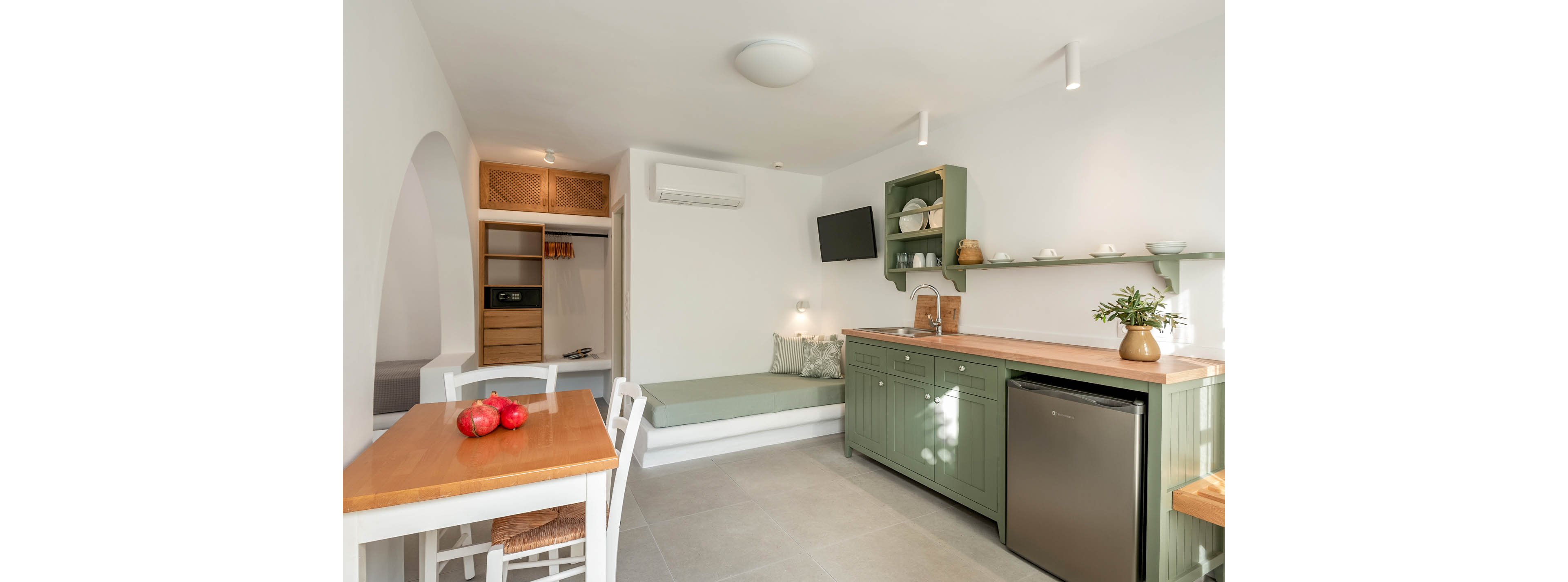 Compact studio interior featuring a small kitchen with sage green cabinets and wooden countertops. A built-in bench with cushions sits beside a wall-mounted television and air conditioner. Open shelving displays dishes while a small refrigerator and sink complete the kitchenette. In the foreground, a wooden dining table holds red apples, and a doorway reveals a simple wardrobe area.