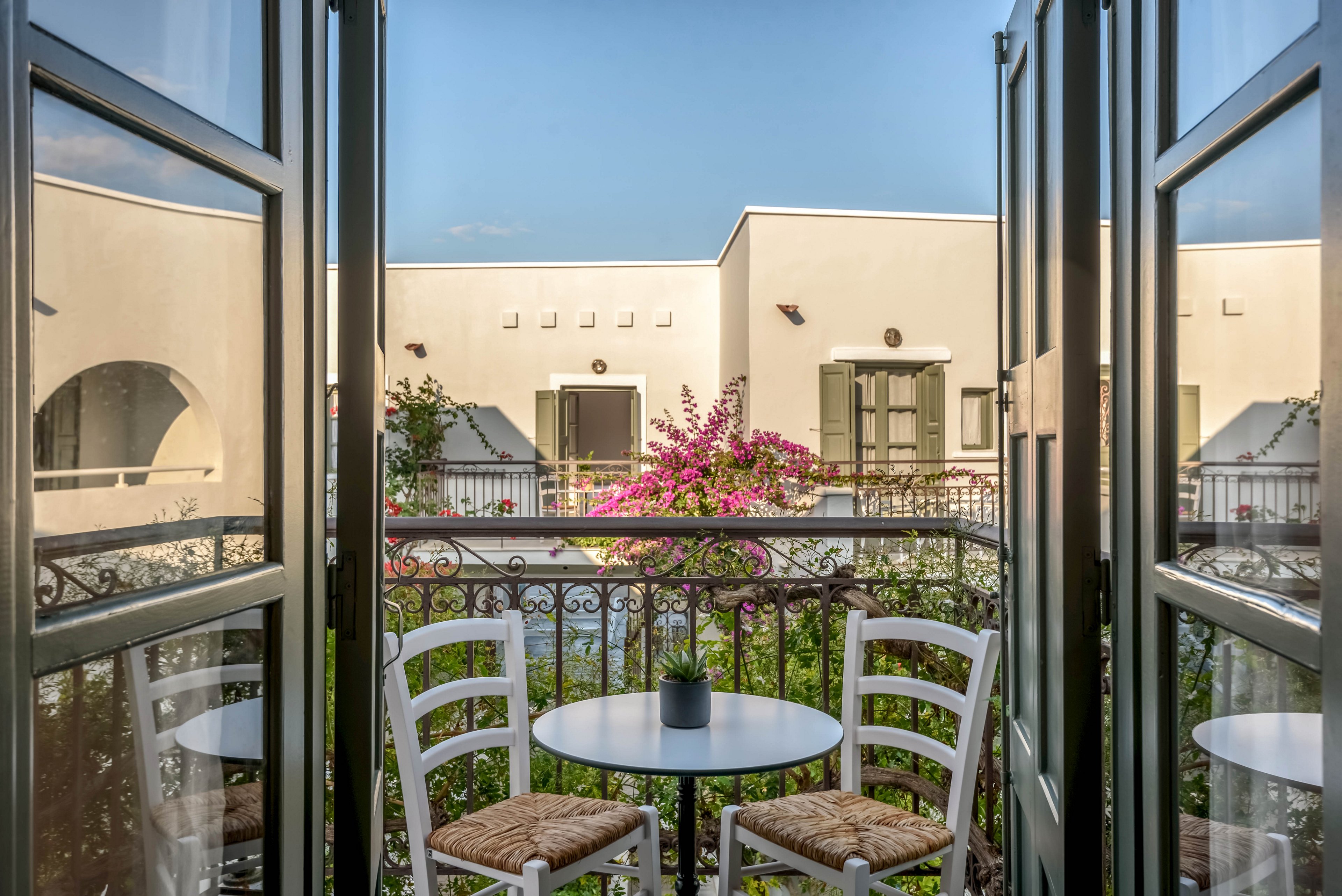 A small balcony with white wooden chairs and a round table is framed by open glass doors. A potted plant sits on the table, overlooking a courtyard of white stucco buildings. Pink bougainvillea flowers add color to the scene, contrasting with the clear blue sky. The setting suggests a peaceful Mediterranean or coastal hotel or residence, ideal for relaxing outdoors.
