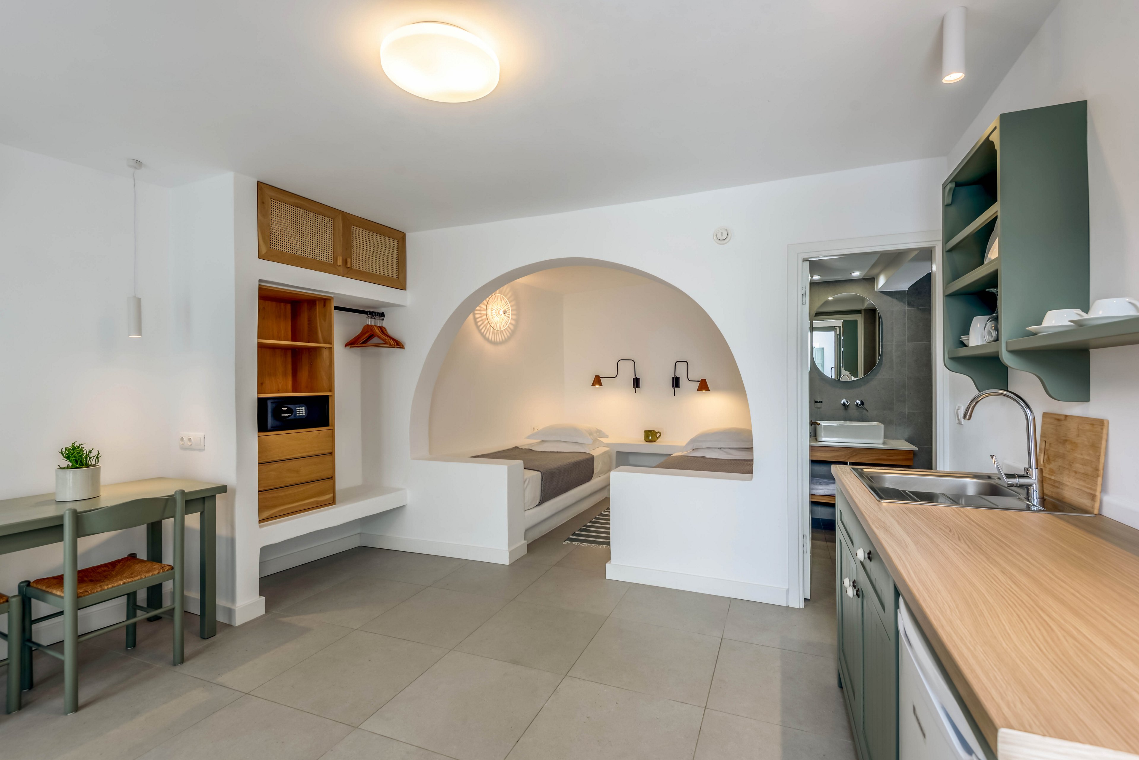 Bright studio apartment interior featuring a built-in arched sleeping nook with twin beds. The space includes a compact kitchenette with green cabinets and wooden countertops on the right. A small dining table with a potted plant and open shelving with hanging space and microwave are visible on the left. Neutral tones, clean lines, and soft lighting create a calm, modern atmosphere.