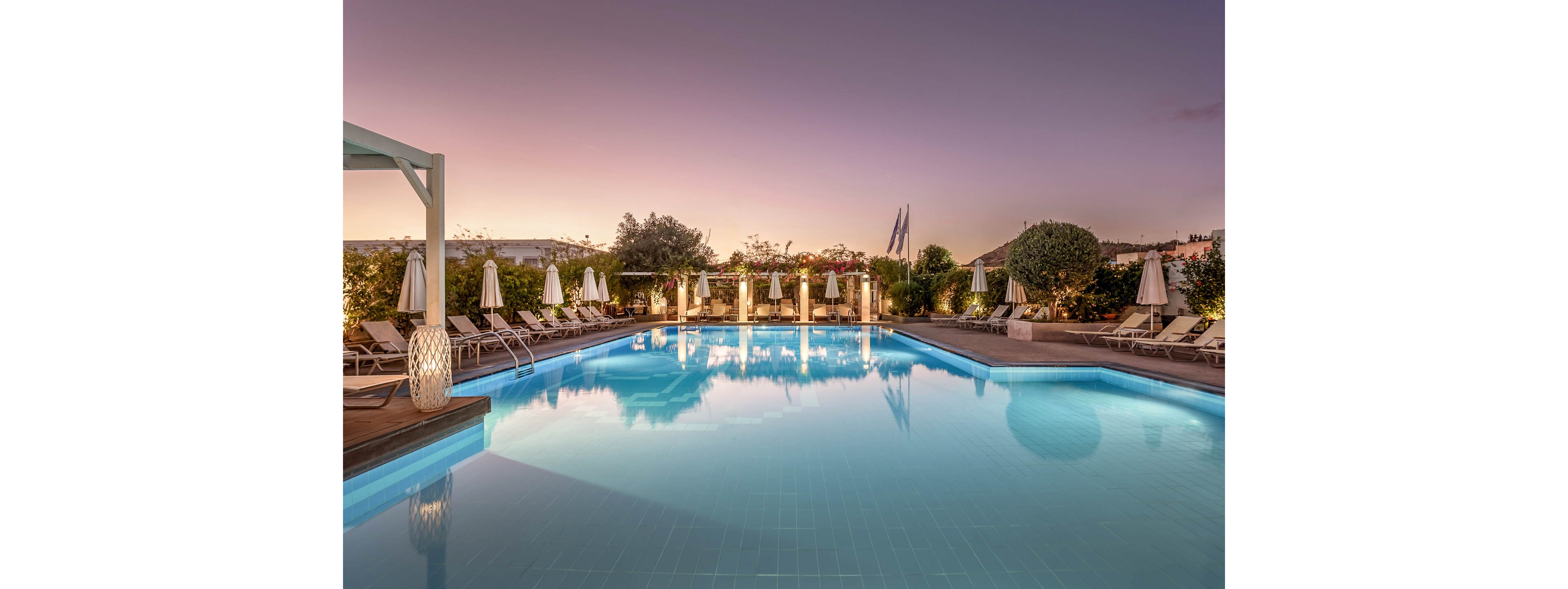 A large outdoor swimming pool reflects the soft colors of a sunset sky. Sun loungers and closed umbrellas line the poolside, suggesting a tranquil resort setting. Warm decorative lights and lush greenery create an inviting, upscale atmosphere. The scene is calm and empty of people, emphasizing relaxation and serenity.