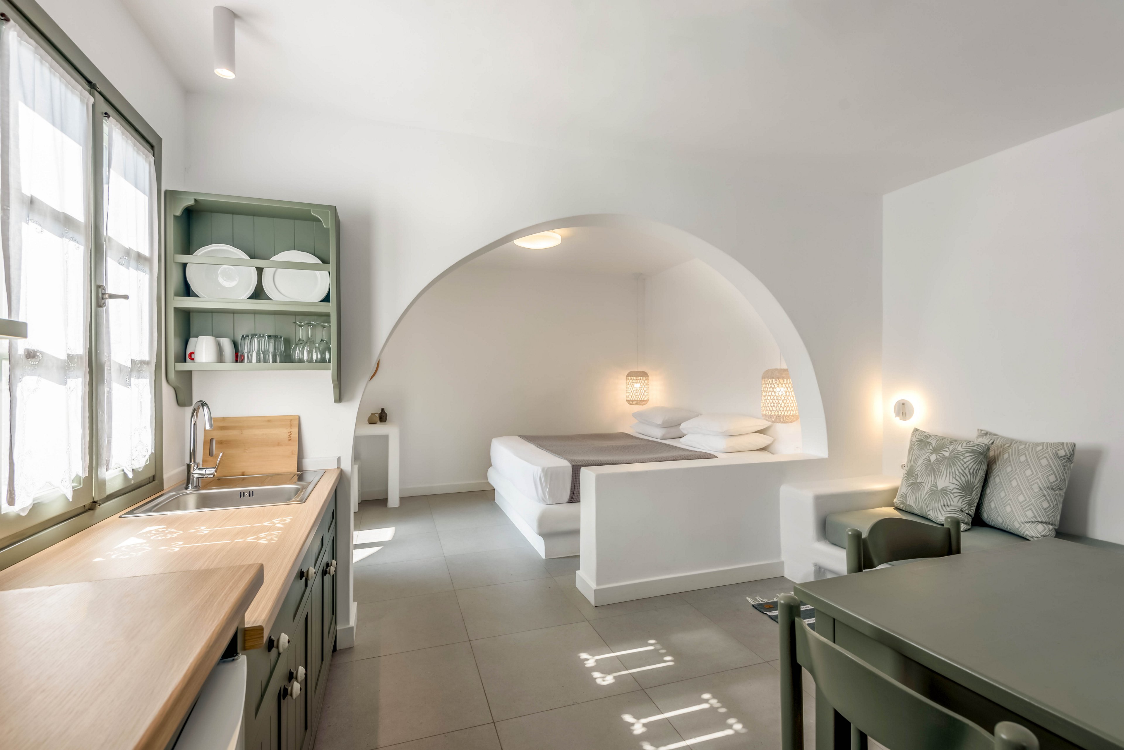 Bright studio interior features a compact kitchen and dining area leading into an arched sleeping nook. The space is decorated in a minimalist style with white walls, light gray flooring, and simple modern furniture. Soft natural light enters through a window above the wooden countertop, illuminating open shelving with dishes and glassware. Neutral textiles and subtle lighting create a calm, contemporary atmosphere suitable for vacation rental or urban living.