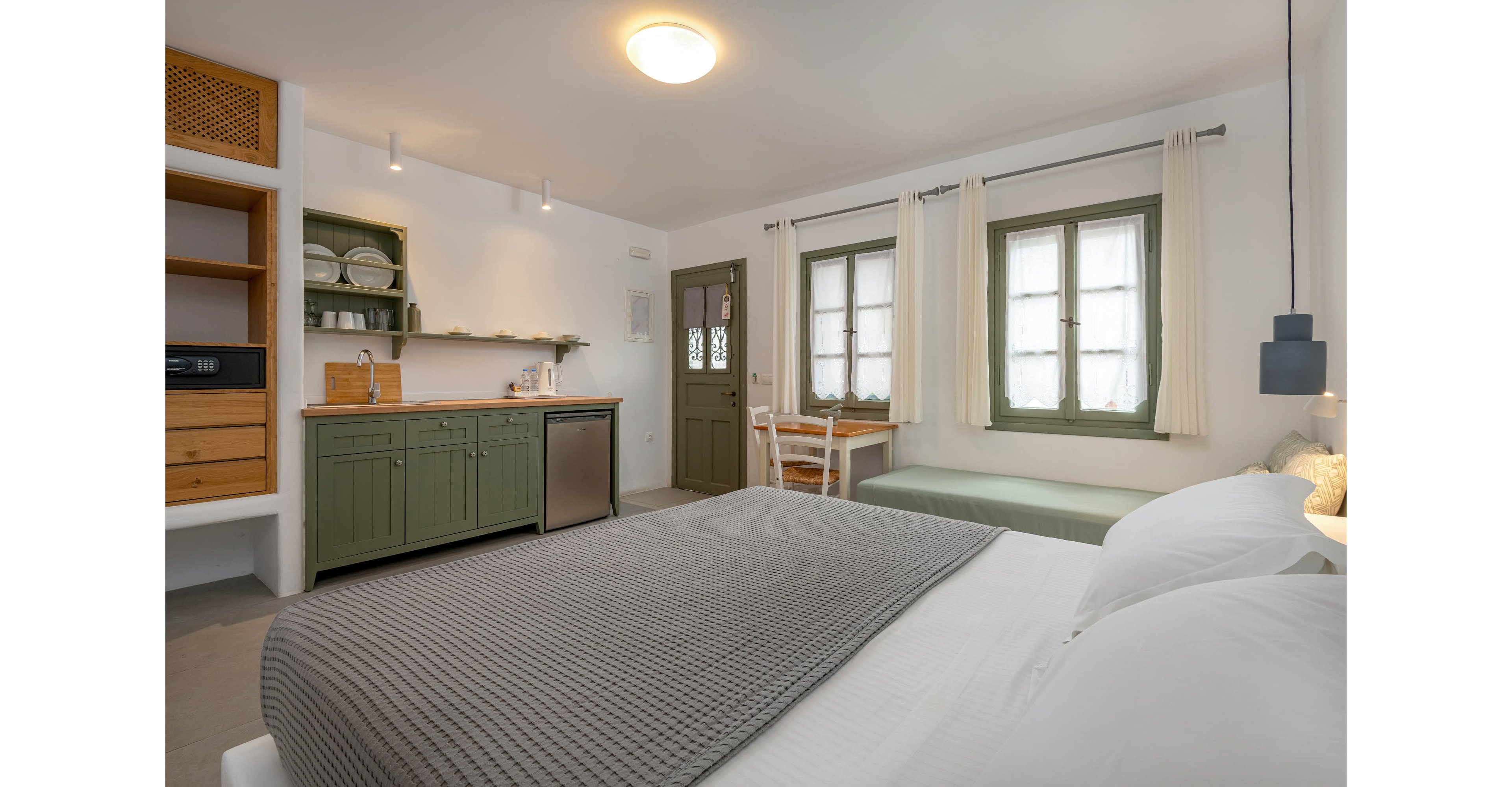 Bright studio room featuring a neatly made double bed with a gray knitted blanket in the foreground. A compact kitchenette with green cabinets, open shelving, and a small refrigerator lines the left wall. Two windows with light curtains and a cushioned bench create a relaxed seating area. Soft, warm lighting and neutral tones give the space a calm, minimalist atmosphere.