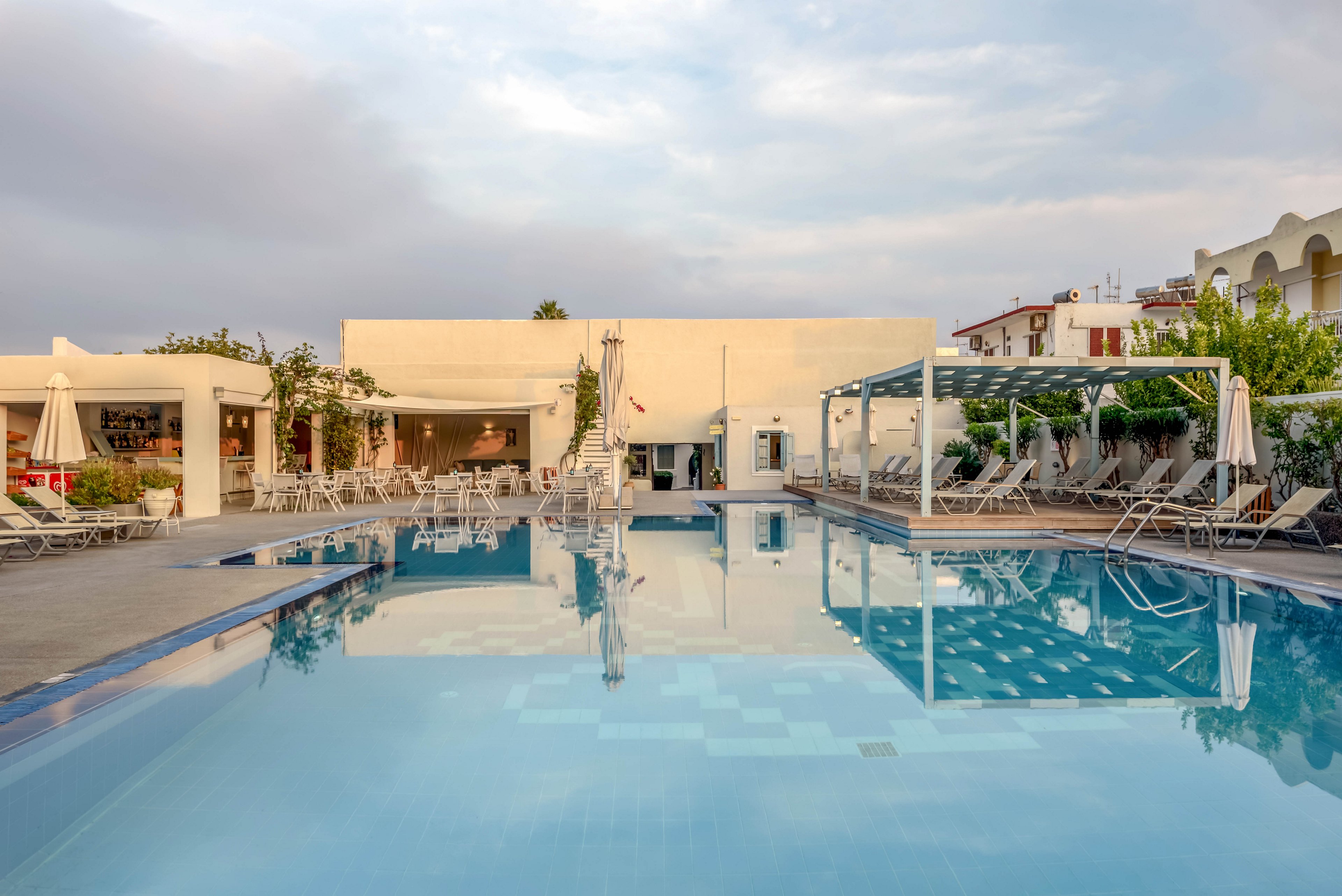A tranquil outdoor swimming pool reflects a modern hotel building under a soft evening sky. Sun loungers and closed umbrellas line the poolside, suggesting a quiet moment between guest activities. The architecture features clean white walls, open terraces, and a shaded pergola area. Lush greenery and neighboring buildings frame the scene, enhancing the relaxed resort atmosphere.