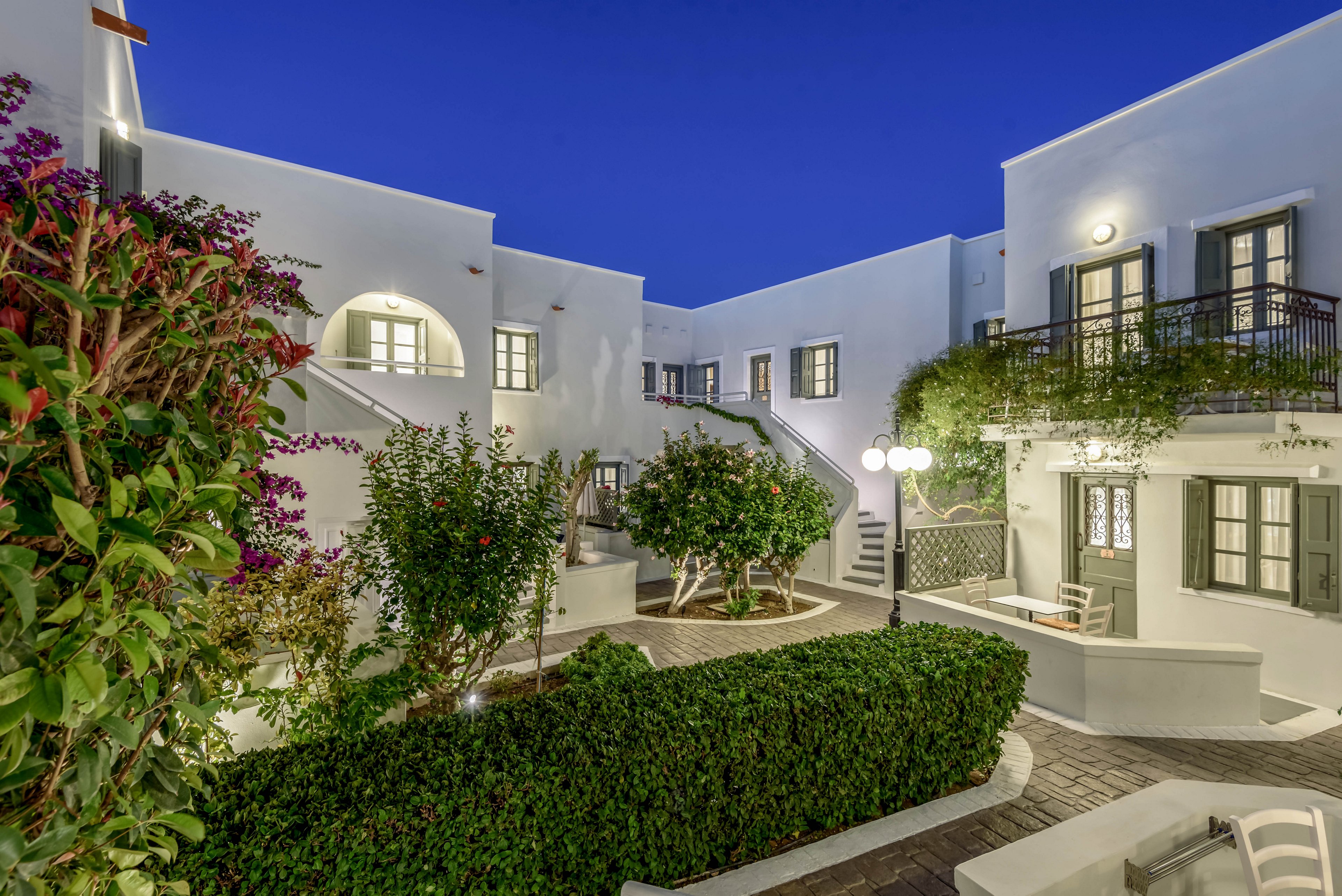 A serene courtyard is surrounded by whitewashed Mediterranean-style buildings under a deep blue evening sky. Neatly trimmed hedges and flowering shrubs frame the stone pathways and staircases. Warm lights from wall lamps and street-style fixtures illuminate doors, windows, and balconies. The scene evokes a peaceful vacation atmosphere in a coastal or island resort setting.