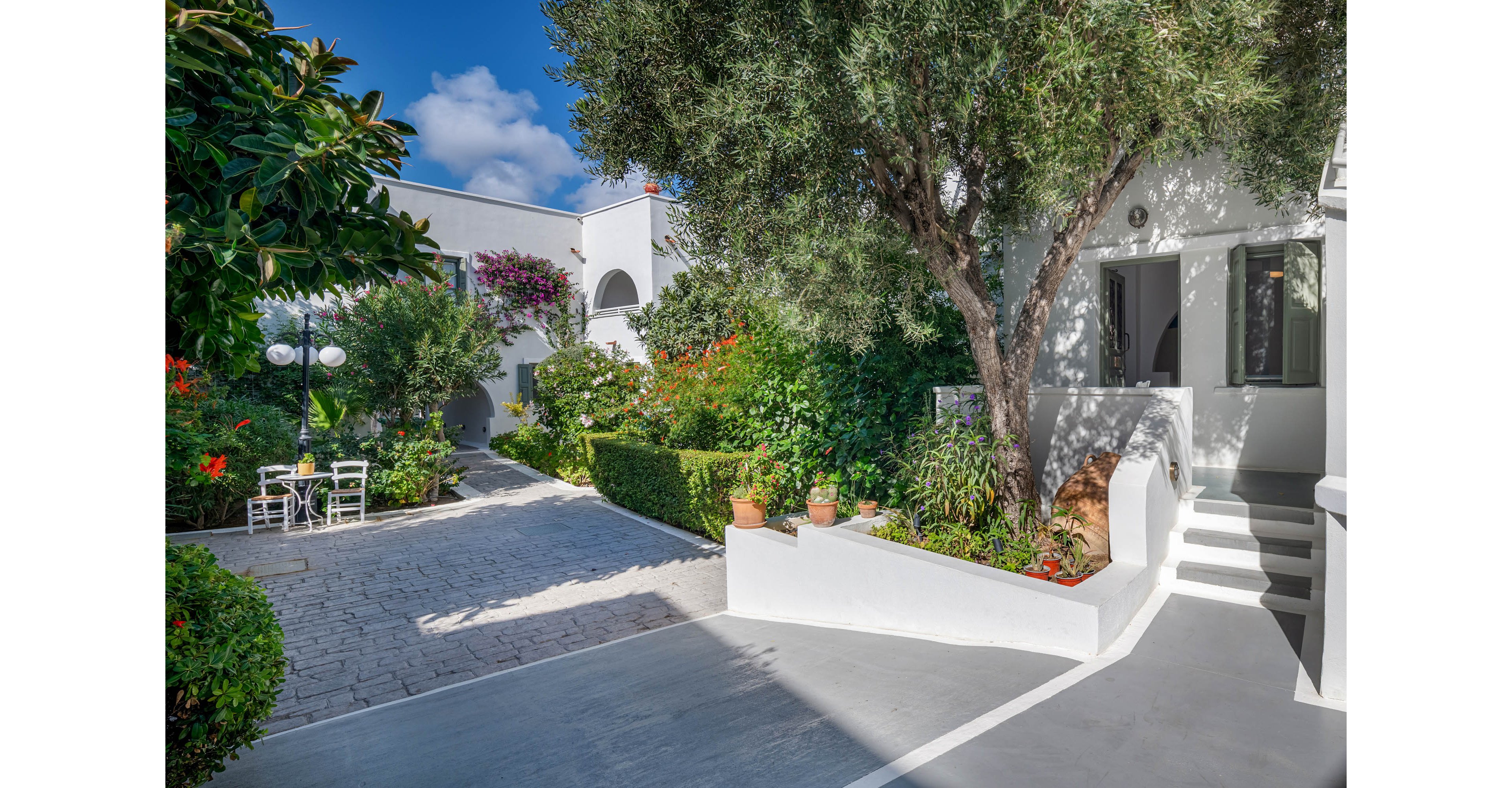 A bright courtyard features white-walled villas surrounded by lush greenery and flowering plants. A stone pathway curves through the garden, leading past trimmed hedges and potted plants. An outdoor seating area with white chairs and a table sits beneath leafy trees. Clear blue sky and strong sunlight create a warm, inviting Mediterranean atmosphere.