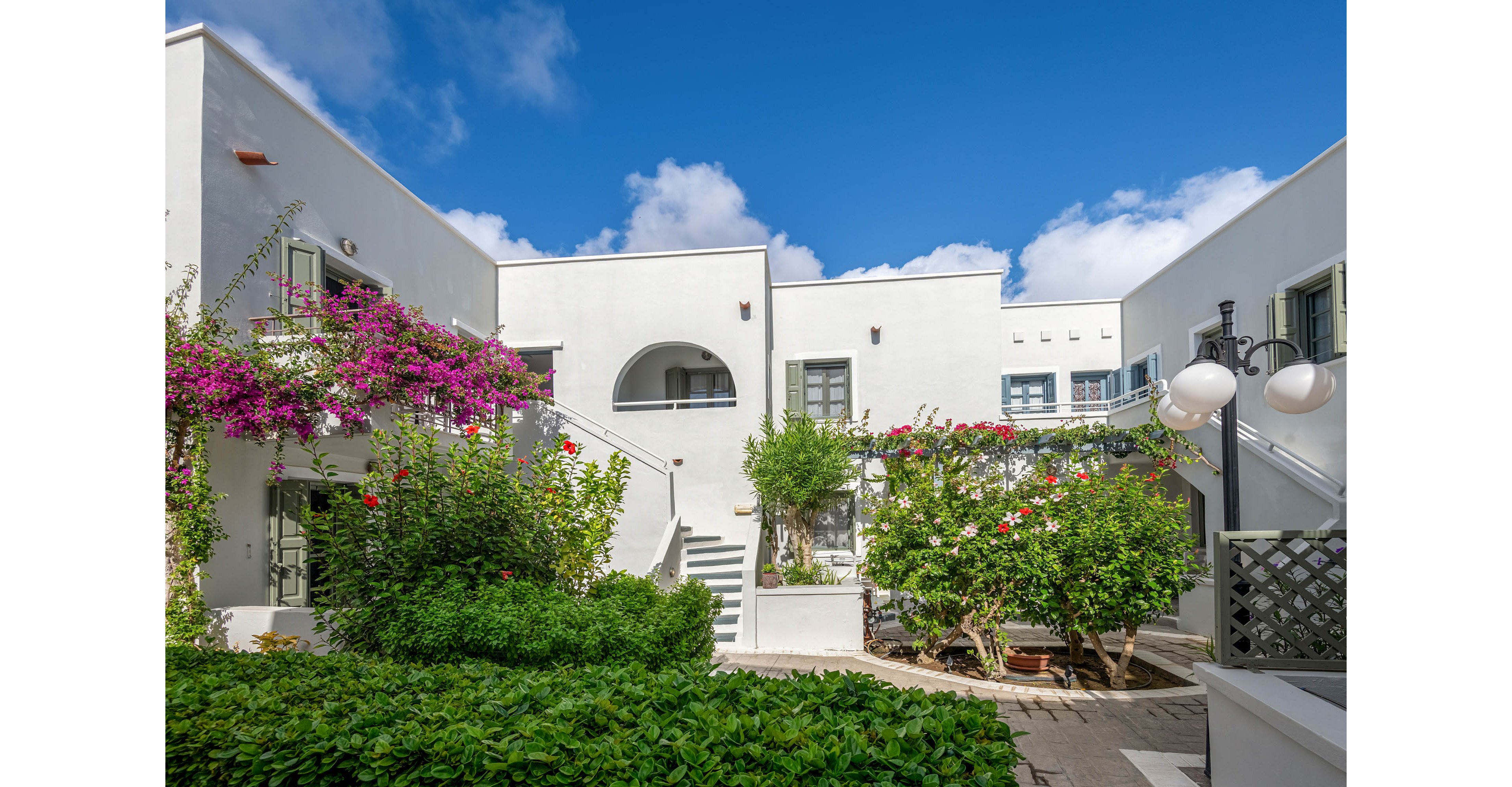 A bright white residential courtyard is framed by low modern buildings under a vivid blue sky. Flowering bushes and climbing vines with pink blossoms soften the clean architectural lines. Green shrubs and potted plants add lush color to the paved courtyard. The scene feels calm and inviting, suggesting a warm Mediterranean or coastal setting.