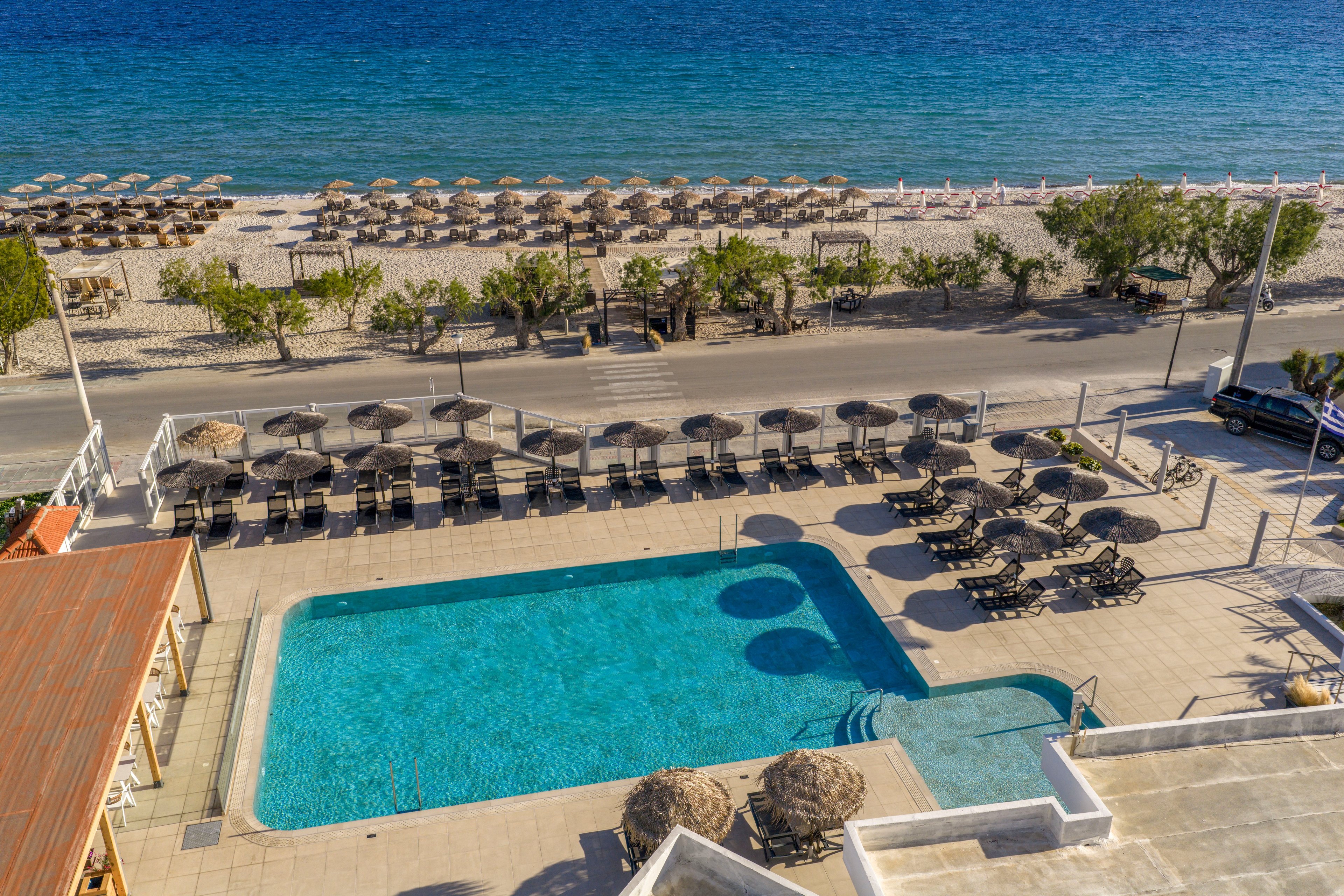 A rectangular outdoor swimming pool is situated in the foreground of a coastal resort. Rows of sun loungers and umbrellas surround the pool, with additional loungers and straw umbrellas arranged along the sandy beach across a small road. Calm blue sea stretches into the distance under clear skies, creating a relaxed vacation atmosphere. The scene is captured from an elevated viewpoint, highlighting the symmetry of the pool area and beach setup.