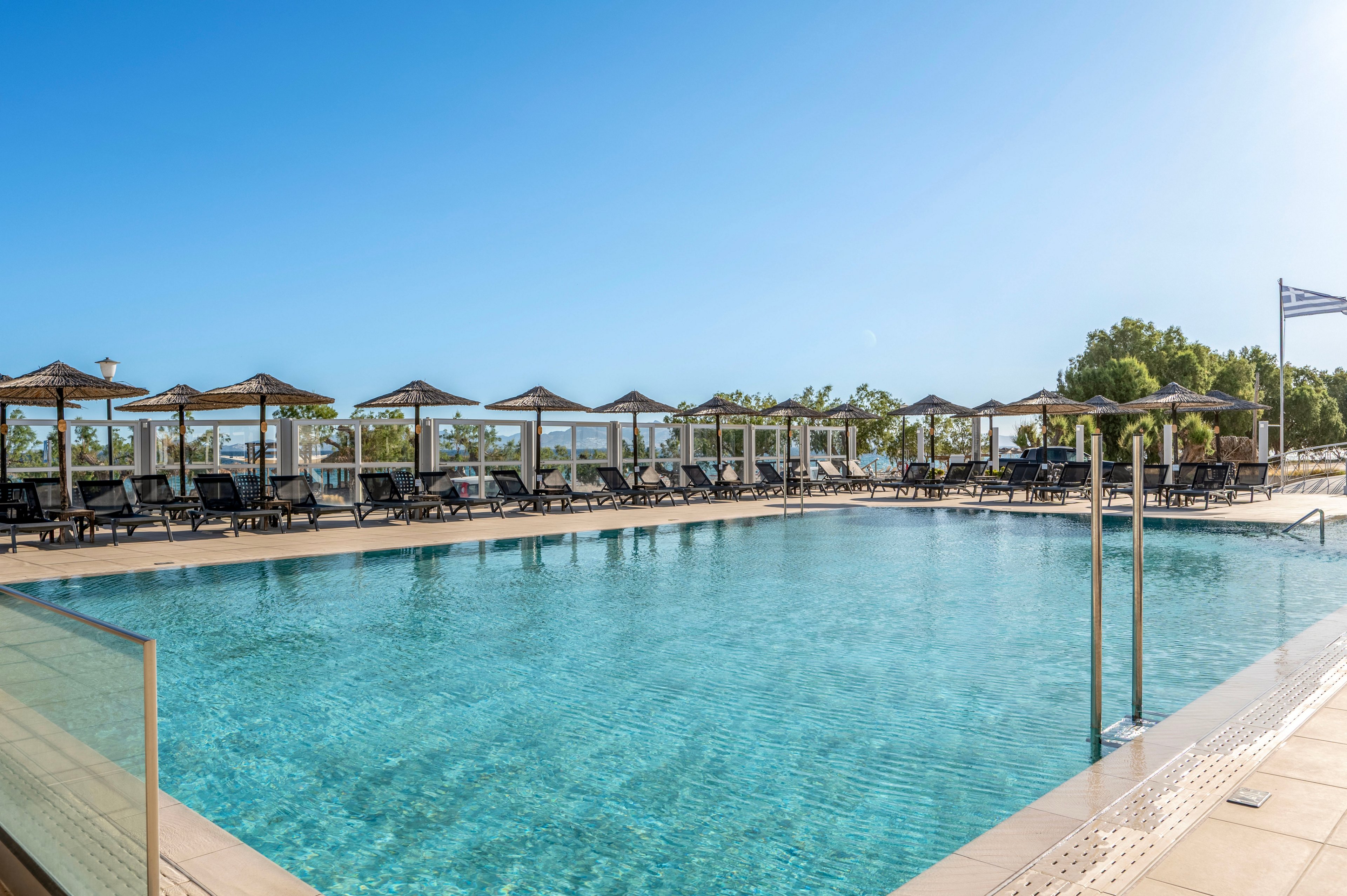 An outdoor swimming pool glistens under a clear blue sky at a resort. Rows of sun loungers and umbrellas line the poolside deck, facing the water. Metal handrails lead into the pool, and trees and greenery are visible in the background. A flag waves on a pole near the edge of the pool area, enhancing the vacation atmosphere.