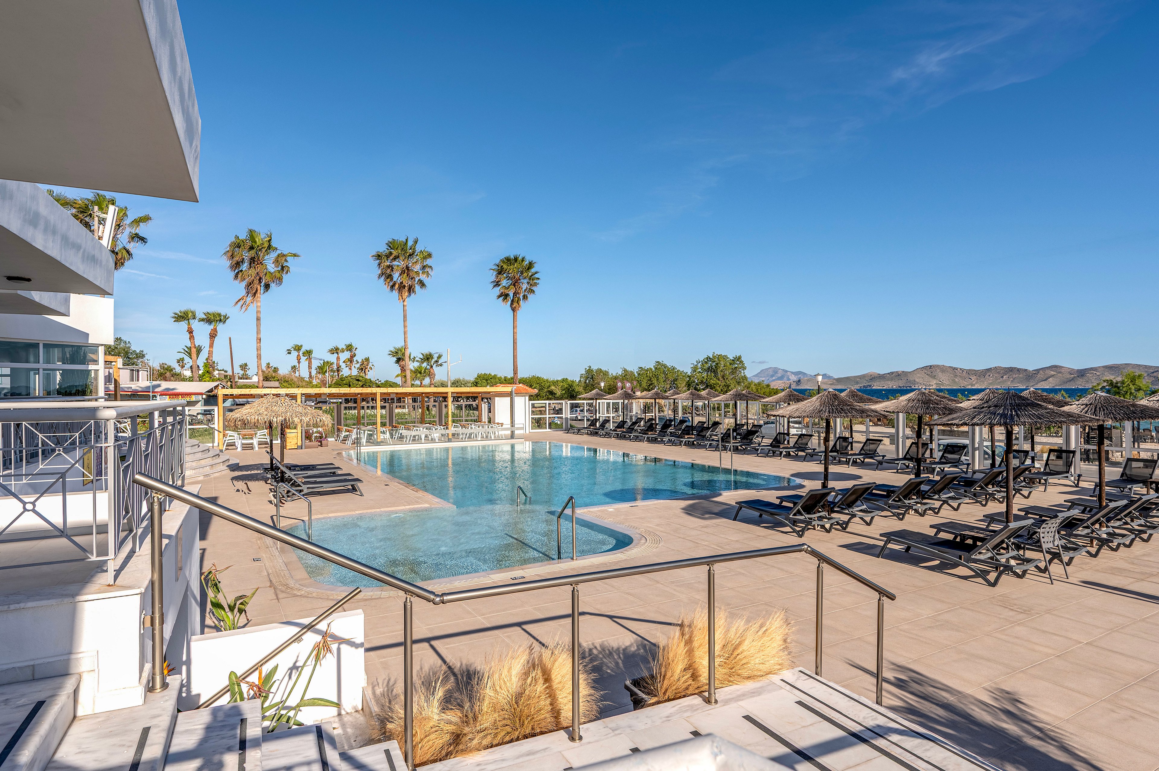 An outdoor resort swimming pool is surrounded by numerous sun loungers and parasols under a clear blue sky. Palm trees line the far side of the pool, adding a tropical feel to the modern terrace setting. Metal railings, tiled flooring, and landscaped grasses frame the pool area, with mountains visible in the distant background. The scene is bright and tranquil, suggesting a relaxing vacation atmosphere.