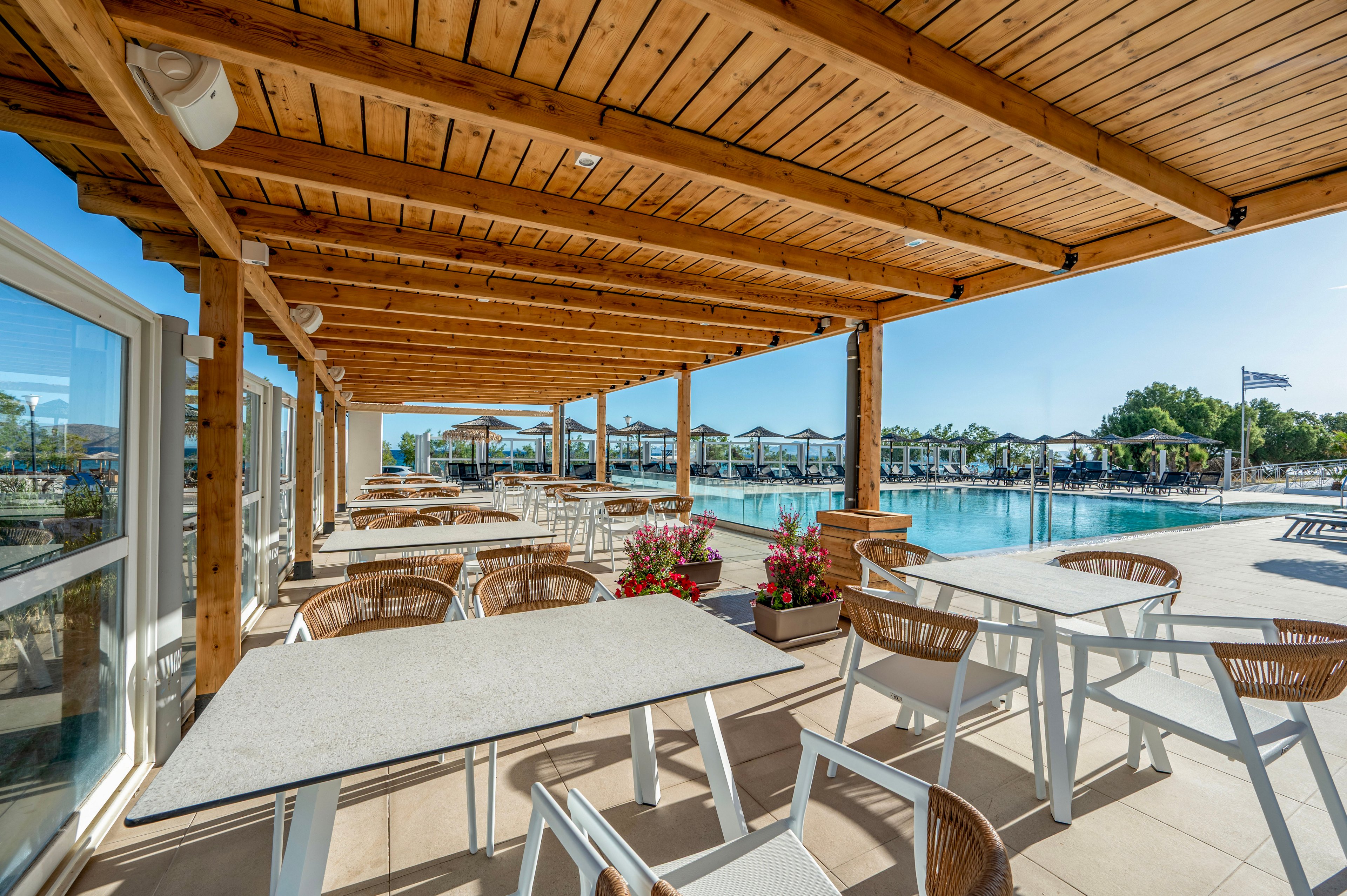 Bright outdoor dining terrace overlooks a large swimming pool under a wooden pergola. White tables and wicker-style chairs are neatly arranged, suggesting a relaxed resort or hotel setting. Flower planters add color near the poolside, while sun loungers and umbrellas line the far edge of the water. Clear blue sky and reflections on the pool create a sunny, vacation atmosphere.