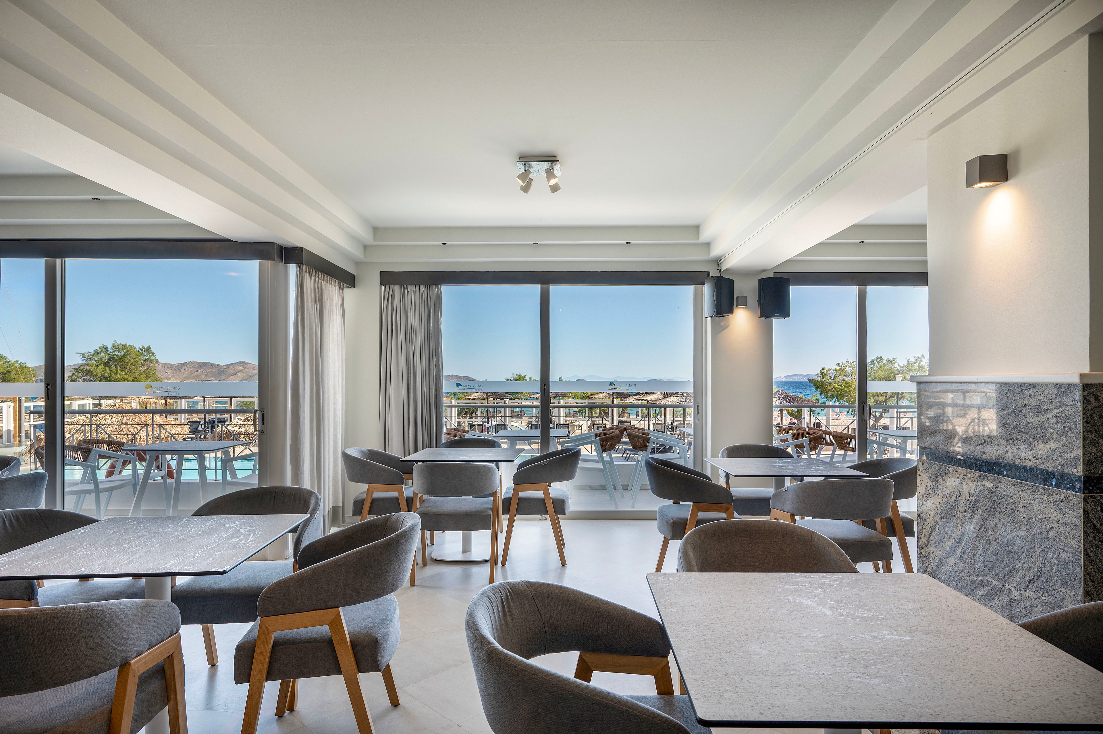 Contemporary indoor seating area with gray upholstered chairs and simple tables faces large floor-to-ceiling windows. Outside, a sunlit pool deck with loungers and umbrellas is visible, along with distant hills and blue sky. Neutral tones and clean lines create a calm, minimalist atmosphere suitable for dining or relaxing. Natural light fills the room, highlighting the spacious, open-plan layout.