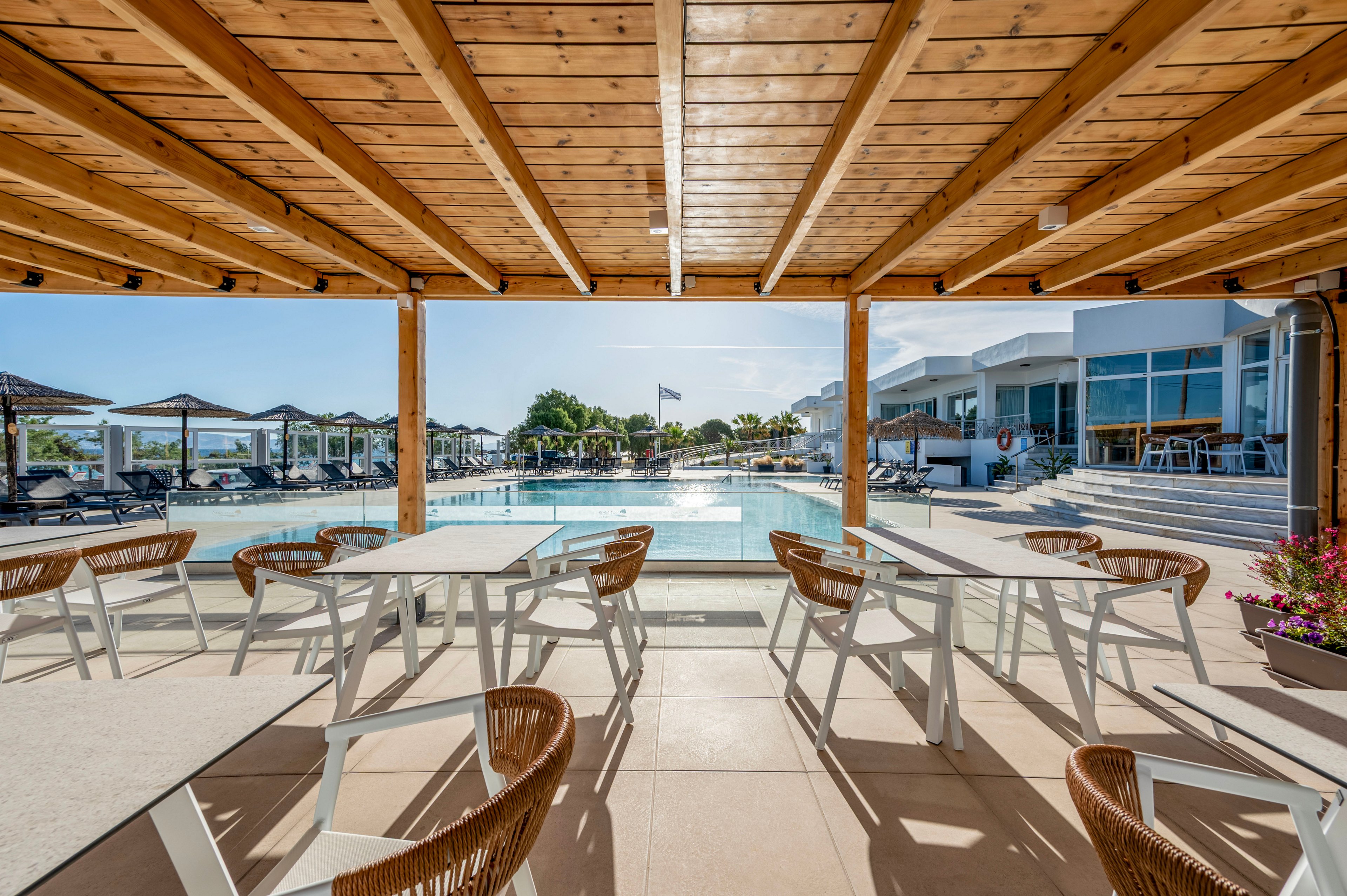 Bright outdoor dining terrace with modern white tables and wicker chairs faces a large resort swimming pool. A wooden pergola ceiling frames the view toward sun loungers and umbrellas around the water. Contemporary white buildings and glass windows line the poolside, suggesting a stylish vacation destination. Clear blue sky and strong sunlight create a relaxed, summery atmosphere.