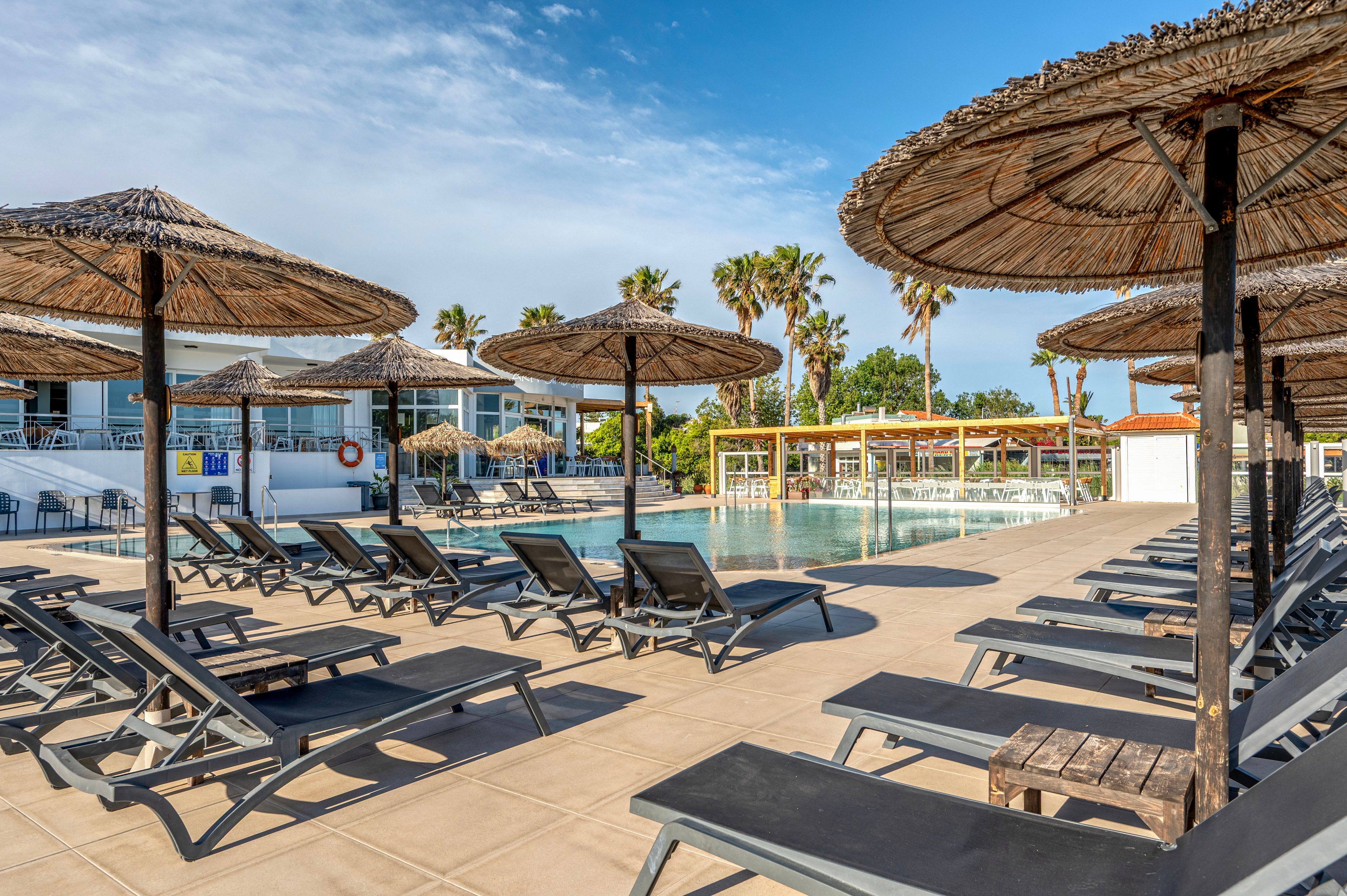A tranquil resort pool area features rows of empty sun loungers under large straw parasols. The swimming pool reflects the clear blue sky and surrounding palm trees, creating a relaxed vacation atmosphere. Modern white buildings and a pool bar structure frame the scene in the background. The setting suggests a sunny, warm day ideal for leisure and outdoor relaxation.