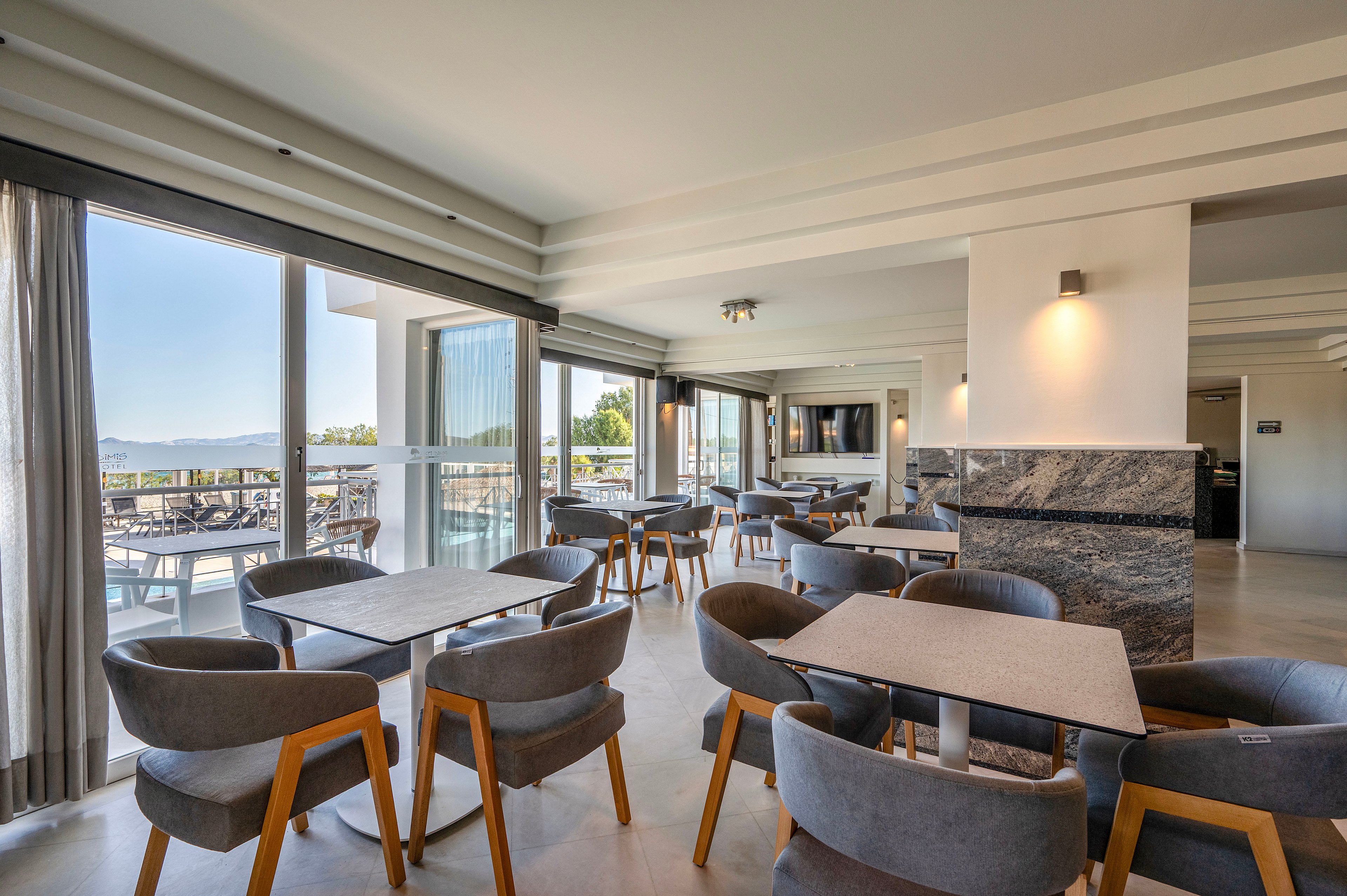 Contemporary lounge area with multiple tables and cushioned chairs is shown in a bright, open interior. Large floor-to-ceiling windows and sliding doors lead to an outdoor terrace with additional seating and scenic views. Neutral tones, marble accents, and minimalist lighting create a clean, modern atmosphere. Sunlight streaming through the windows enhances the airy, relaxed feel of the space.