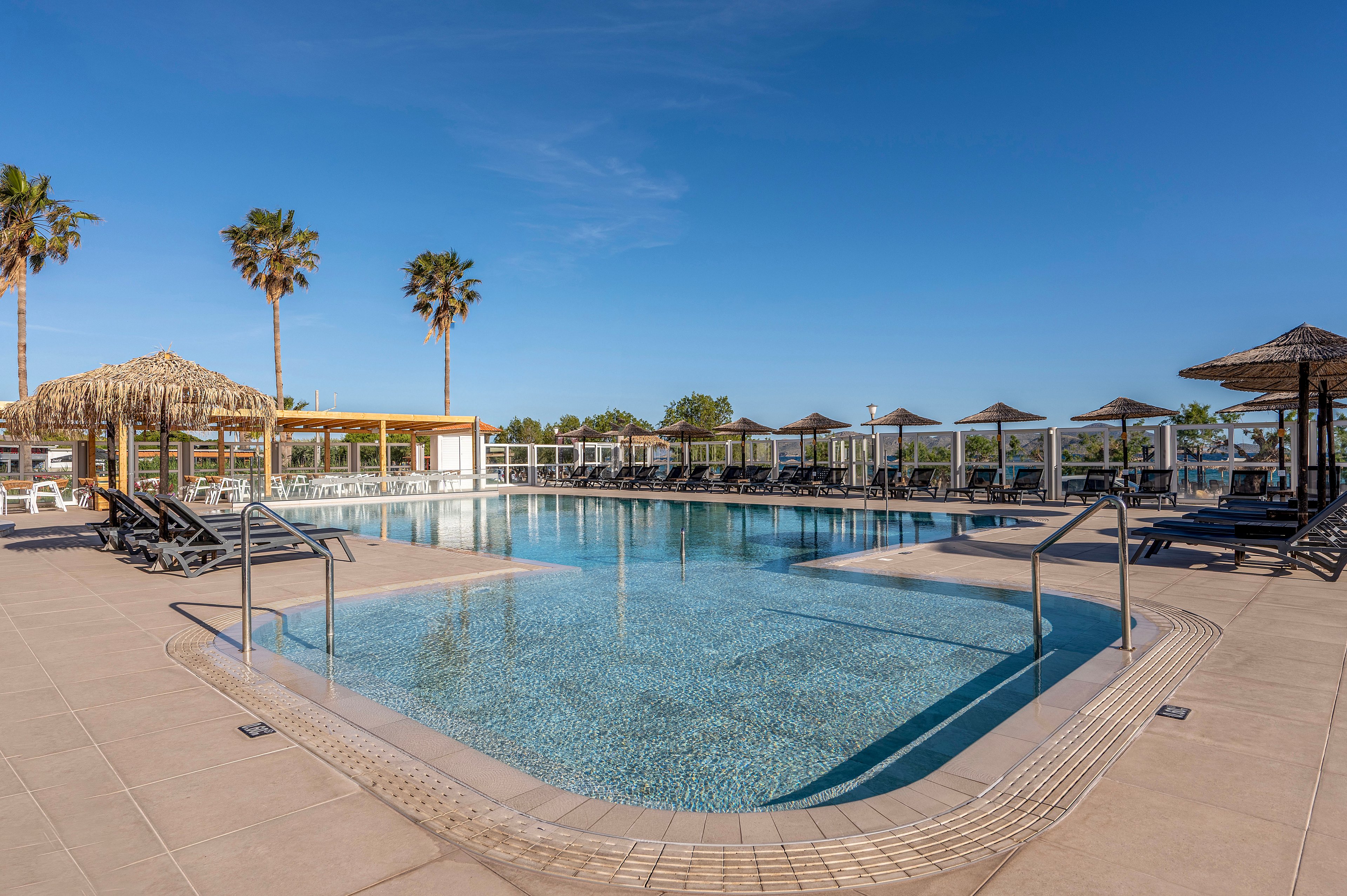 A large, clear swimming pool sits at the center of a modern resort under a bright blue sky. Sun loungers line the tiled deck, with thatched umbrellas and cabanas providing shaded seating around the water. Tall palm trees rise in the background, enhancing the tropical vacation atmosphere. Stainless steel handrails and clean geometric lines give the pool area a contemporary, well-maintained look.
