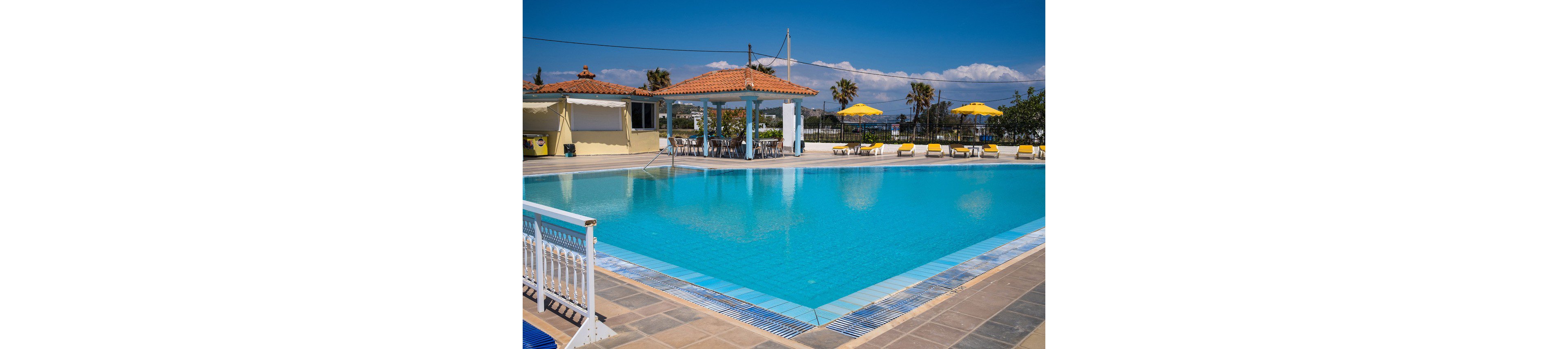 A large outdoor swimming pool glistens under a clear blue sky at a coastal resort. Yellow sun loungers and umbrellas line the far side of the pool, suggesting a relaxing holiday atmosphere. A small building with a tiled roof and shaded terrace sits beside the water, with railings and steps leading into the pool. Palm trees and distant sea views complete the bright, summery setting.