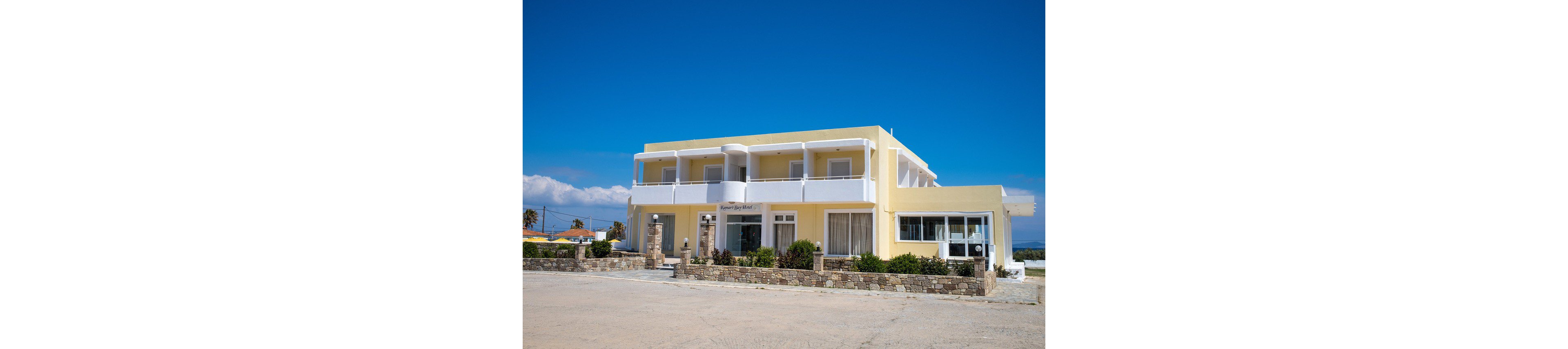 A two-story yellow hotel building stands under a clear blue sky. The structure features multiple balconies with white railings and large windows on both floors. A stone wall with small shrubs and plants borders the front of the property. The surrounding area appears quiet and open, with a paved foreground and distant landscape in the background.