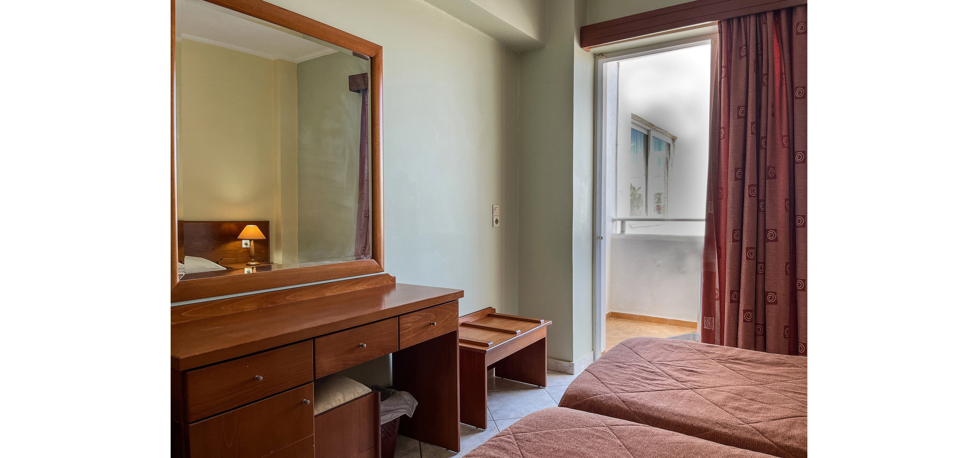 A modest hotel room features twin beds with matching brown bedspreads. A wooden dresser with a large mirror and multiple drawers stands against the wall, reflecting a lit bedside lamp. Heavy curtains frame a doorway leading to a small balcony area with tiled flooring. The overall atmosphere is simple, clean, and functional, suitable for budget-friendly accommodation.