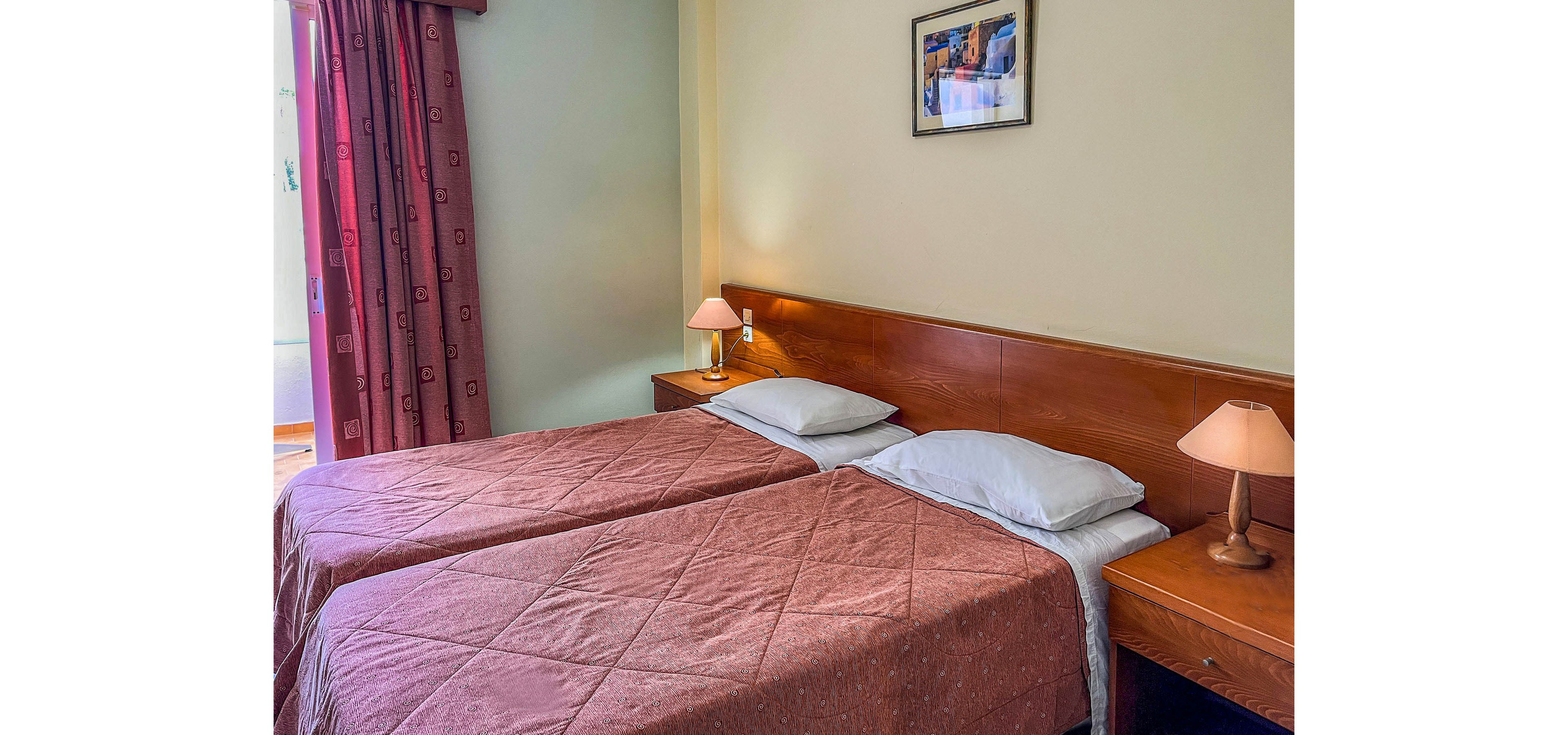 The image shows a simple hotel room with two twin beds placed side by side, each covered with a quilted brown bedspread. A wooden headboard runs behind both beds, with matching wooden nightstands on either side. Two warm-toned bedside lamps provide soft lighting, and a framed picture hangs on the wall above the headboard. A window with patterned curtains is partially visible, adding natural light to the calm, tidy space.