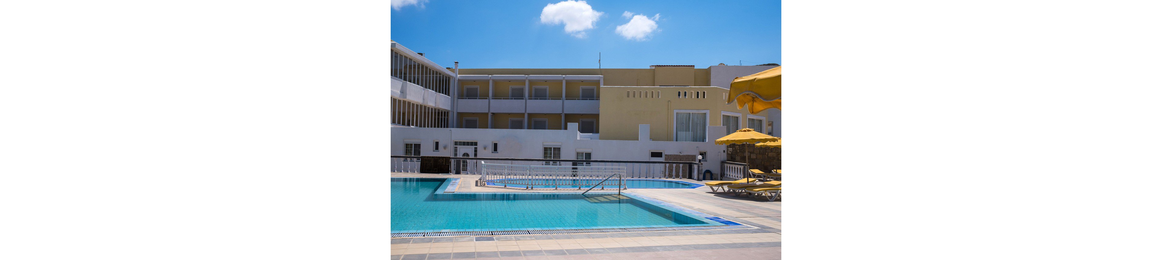 An outdoor swimming pool sits in the courtyard of a modern hotel building under a bright blue sky. The multi-story structure features balconies and large windows overlooking the water. Yellow sun loungers and umbrellas line the tiled pool deck, suggesting a relaxing vacation setting. Clear turquoise water and clean architectural lines create a calm, inviting atmosphere.