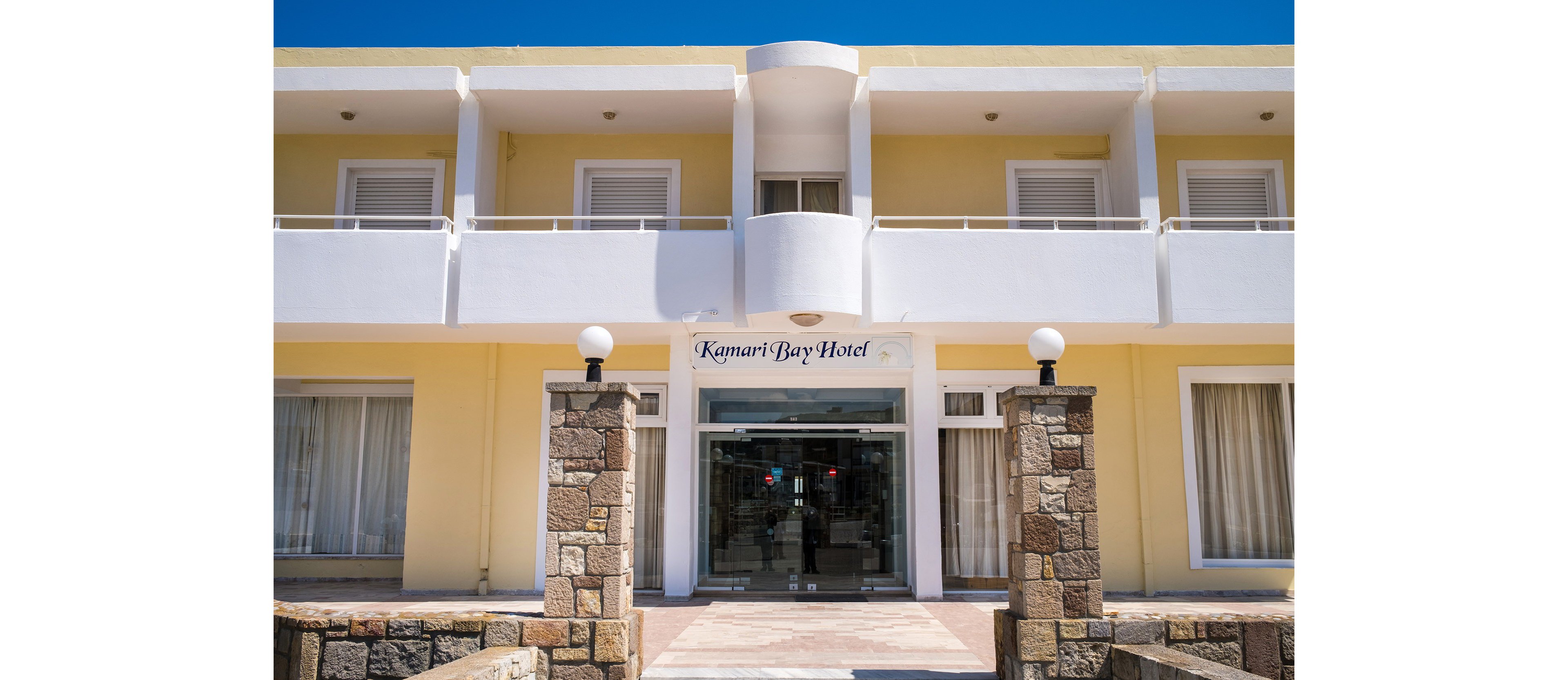 Front view of the Kamari Bay Hotel building with a clear blue sky above. The image shows a symmetrical facade with balconies, large windows, and a central glass entrance. Stone pillars with spherical lights frame the pathway leading to the doorway. The overall scene suggests a bright, sunny day at a coastal or resort destination.
