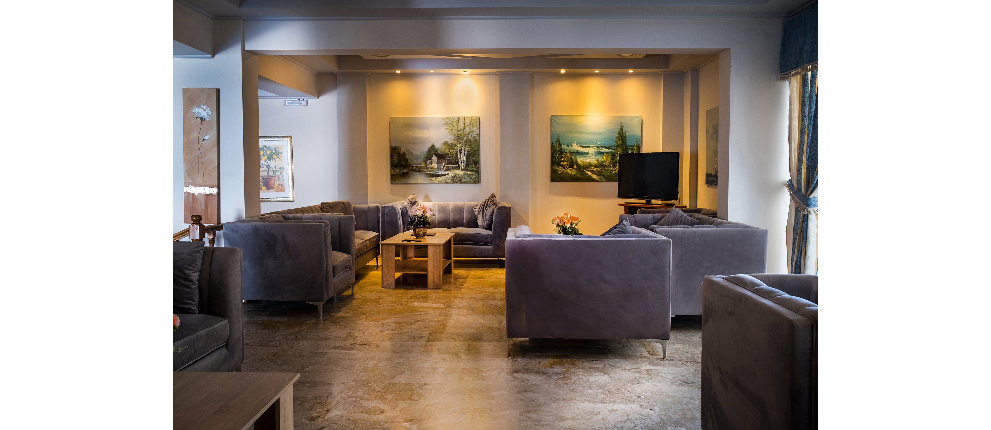 A warmly lit lounge area features multiple gray velvet sofas arranged around low wooden coffee tables. The space appears to be part of a hotel or upscale residence, with polished stone flooring and soft ambient lighting from ceiling spotlights. Large landscape paintings decorate the walls, and a television stands in the corner near a window with draped curtains. Small flower arrangements on the tables add a touch of color to the neutral interior.