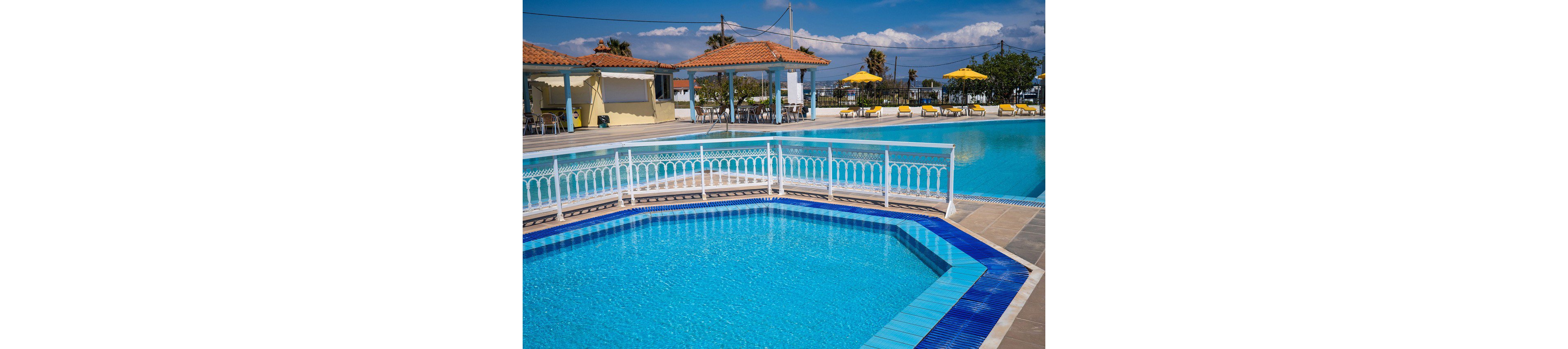 An outdoor resort swimming pool glistens under bright sunlight, featuring a smaller octagonal pool in the foreground. White railings and blue tile accents frame the clear turquoise water. In the background, sun loungers with yellow umbrellas line the poolside near open-air pavilions with terracotta roofs. The scene suggests a warm, relaxing vacation atmosphere at a coastal or holiday destination.