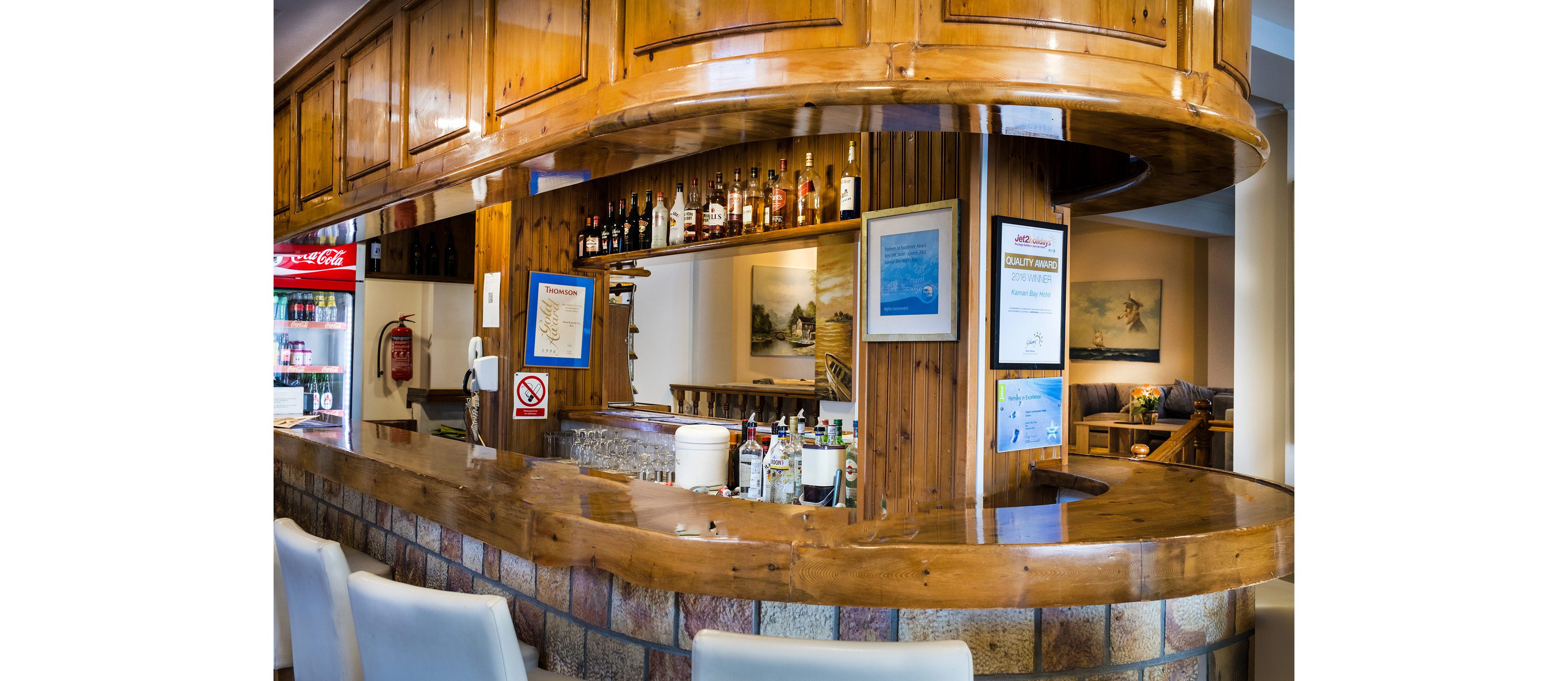 A curved wooden bar counter is stocked with various liquor bottles and bar equipment. The setting appears to be a cozy hotel or lounge bar with framed pictures and certificates on the wall. A refrigerator with drinks is visible to the side, along with a fire extinguisher and a no smoking sign. Warm lighting and wood paneling create an inviting, relaxed atmosphere for guests.