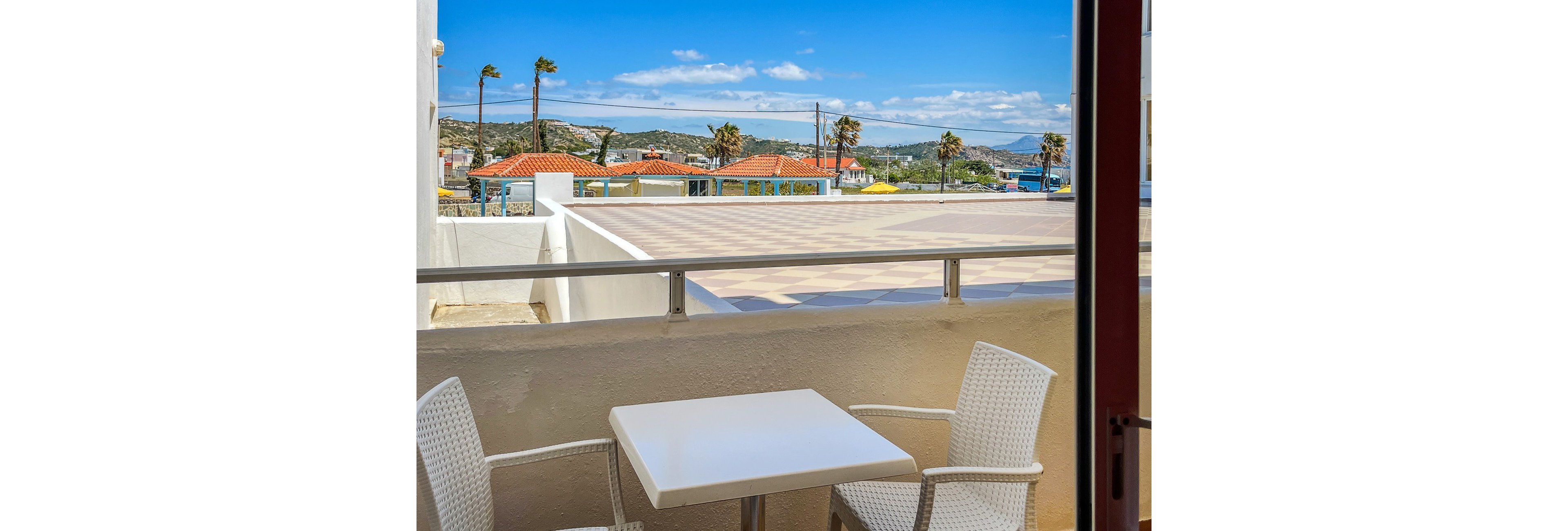 A small balcony features a white square table and two white wicker chairs. Beyond the low wall and metal railing, there is an open terrace area leading to a coastal town. Red-tiled roofs, palm trees, and distant hills are visible under a bright blue sky with scattered clouds. The scene suggests a warm, sunny day in a seaside or Mediterranean setting.