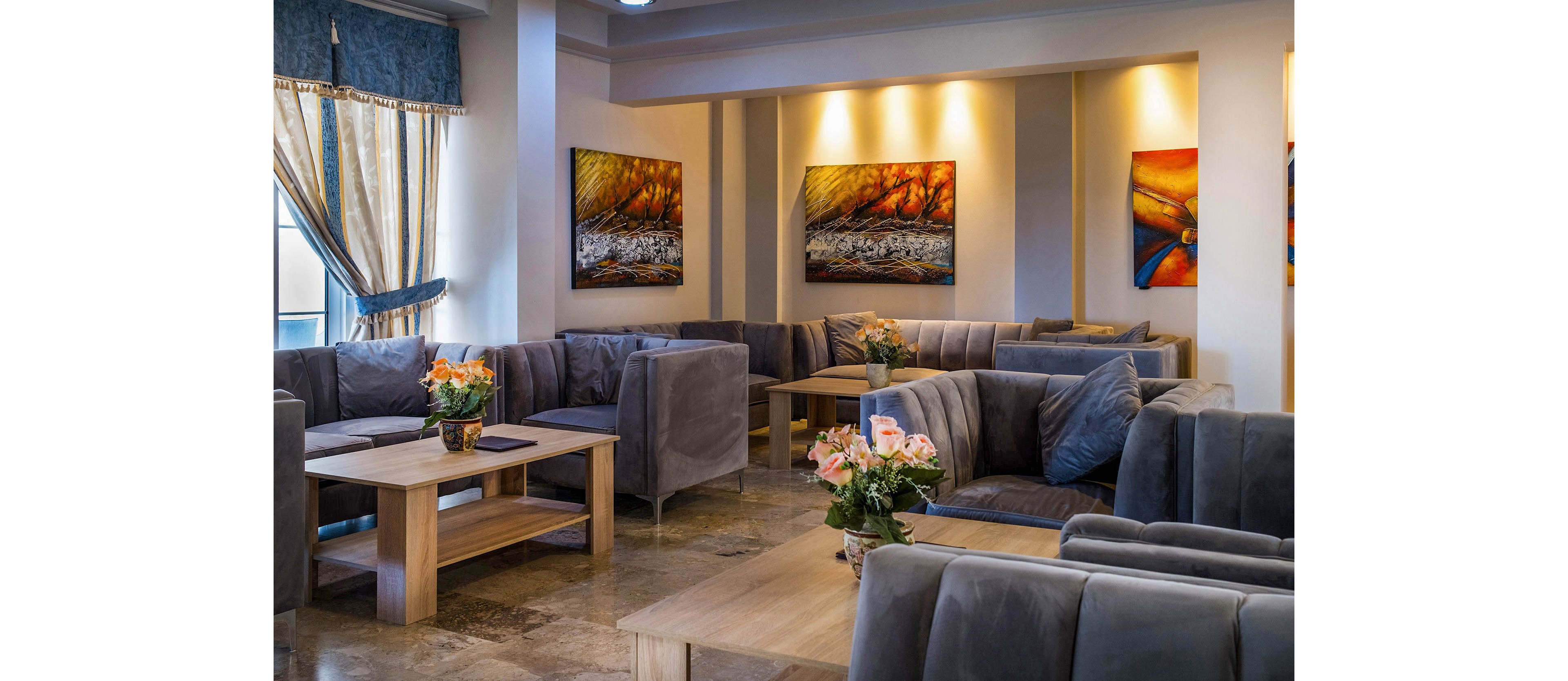 A contemporary lounge area features multiple gray sectional sofas arranged around light wooden coffee tables. Colorful abstract paintings hang on the warmly lit walls, adding contrast to the neutral furniture. Vases of fresh flowers decorate the tables, bringing a touch of color and softness to the space. Large windows with draped curtains allow natural light to complement the ambient interior lighting.