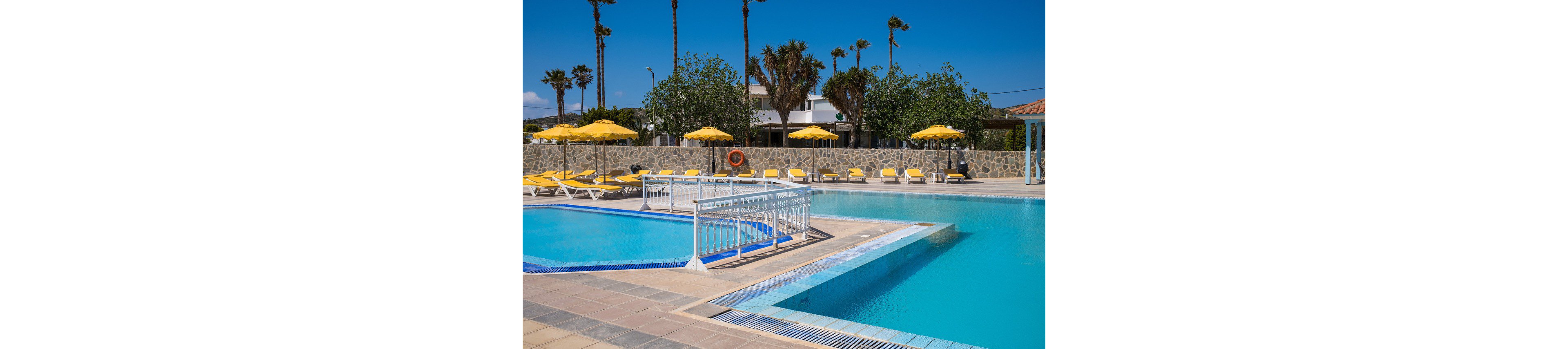 Outdoor resort swimming pools are shown under a clear blue sky. A larger main pool is connected to a smaller shallow pool by a white railing bridge. Yellow sun loungers and matching umbrellas line the pool deck, creating a bright and inviting atmosphere. Palm trees and a stone wall with buildings in the background suggest a warm vacation destination.