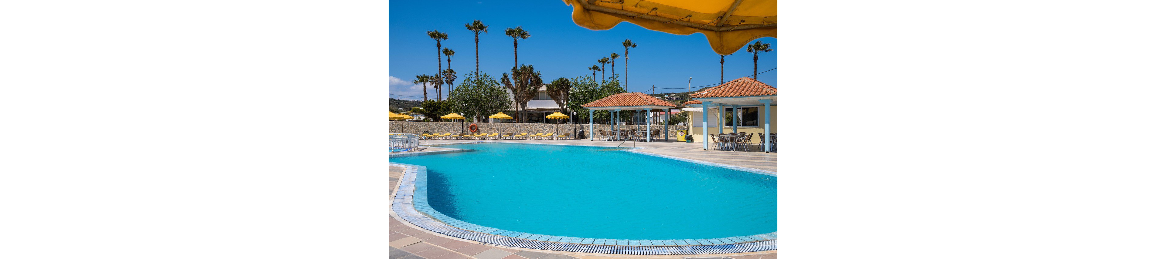 Large outdoor swimming pool at a sunny resort is surrounded by tiled deck and sun loungers. Yellow umbrellas and small pavilion buildings with red-tiled roofs frame the pool area. Tall palm trees and a clear blue sky create a tropical vacation atmosphere. The scene appears calm and inviting, with no people visible.