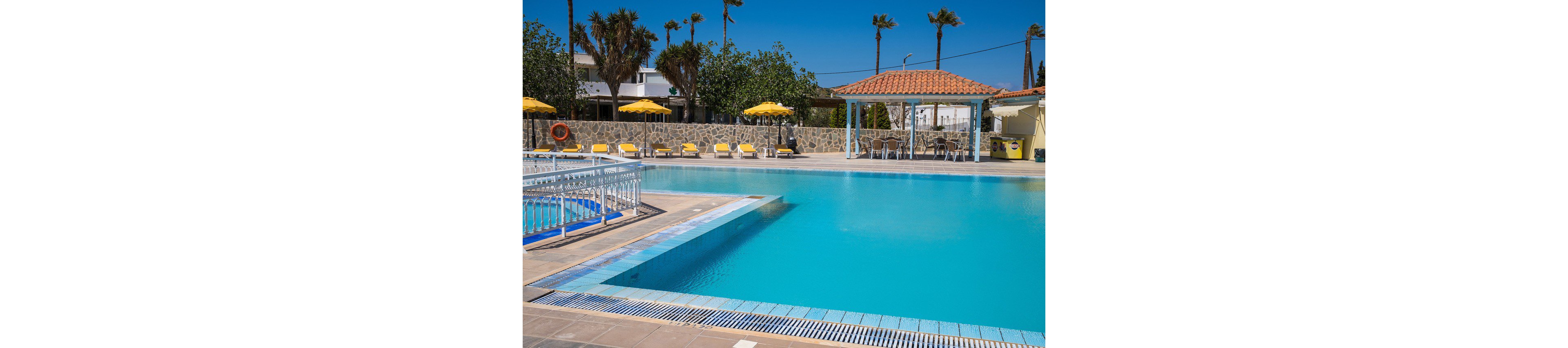 An outdoor swimming pool glistens under a clear blue sky at a resort. Yellow sun loungers and umbrellas line the poolside terrace, suggesting a relaxing vacation setting. A tiled pool bar with a red roof stands nearby, framed by palm trees and a stone wall. The scene feels bright and tranquil, emphasizing leisure and warm weather.
